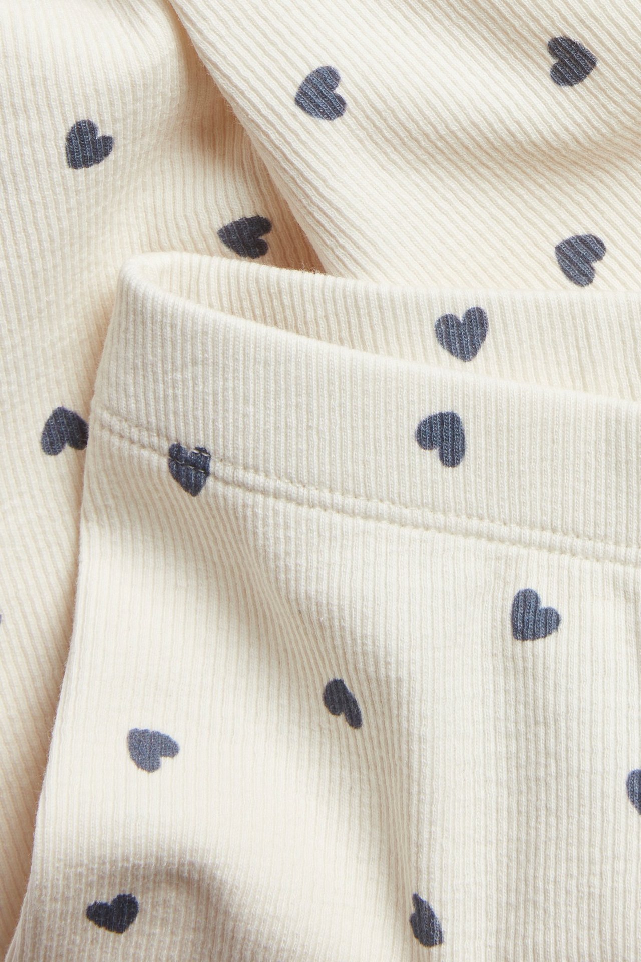 Lähikuva luonnonvalkoisista ribatuista leggingseistä, tummanharmaa sydänkuvio ja joustava vyötärö.