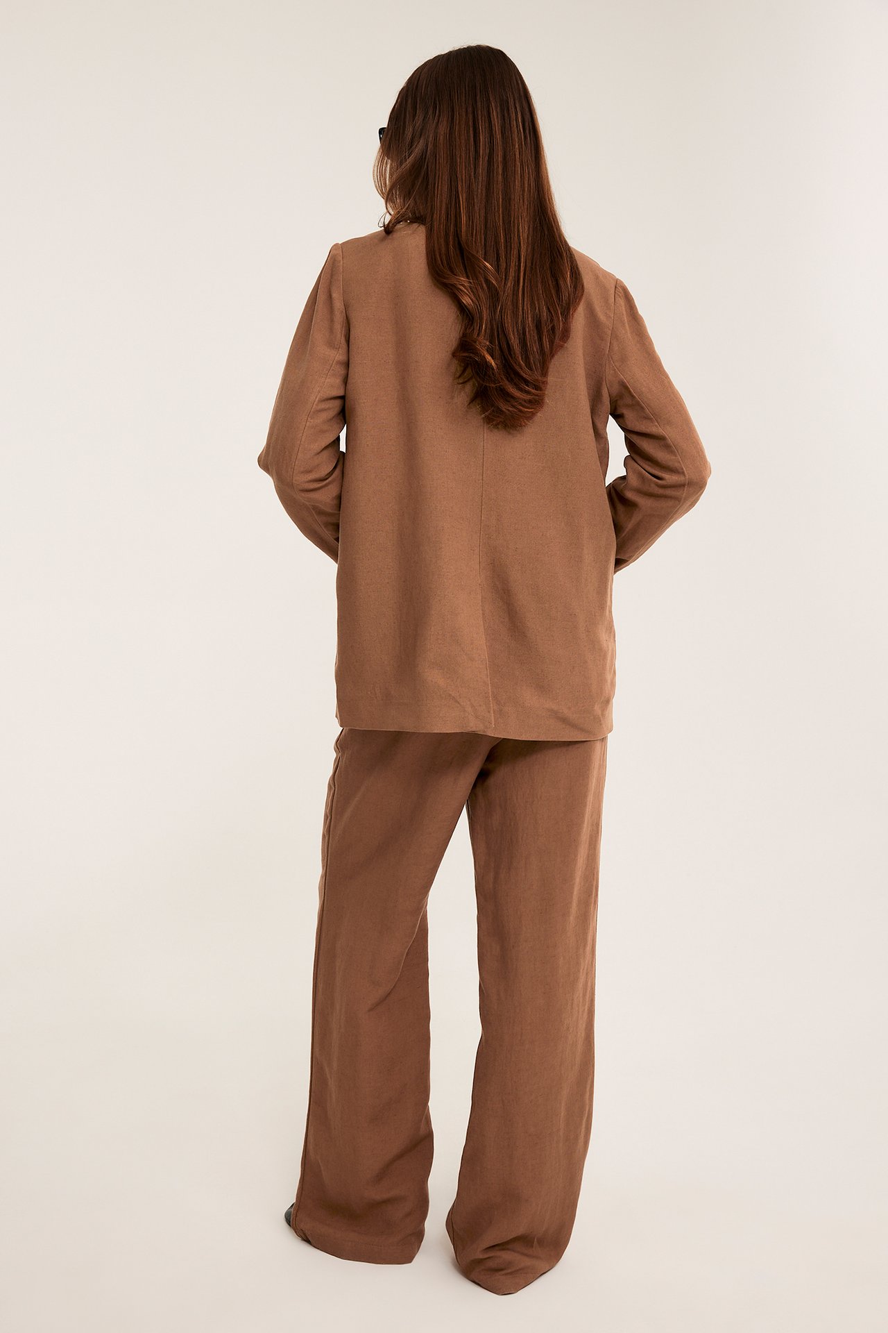 Widok z tyłu: kobieta w luźnych brązowych lnianych spodniach i dopasowanej marynarce z mieszanki lnu.