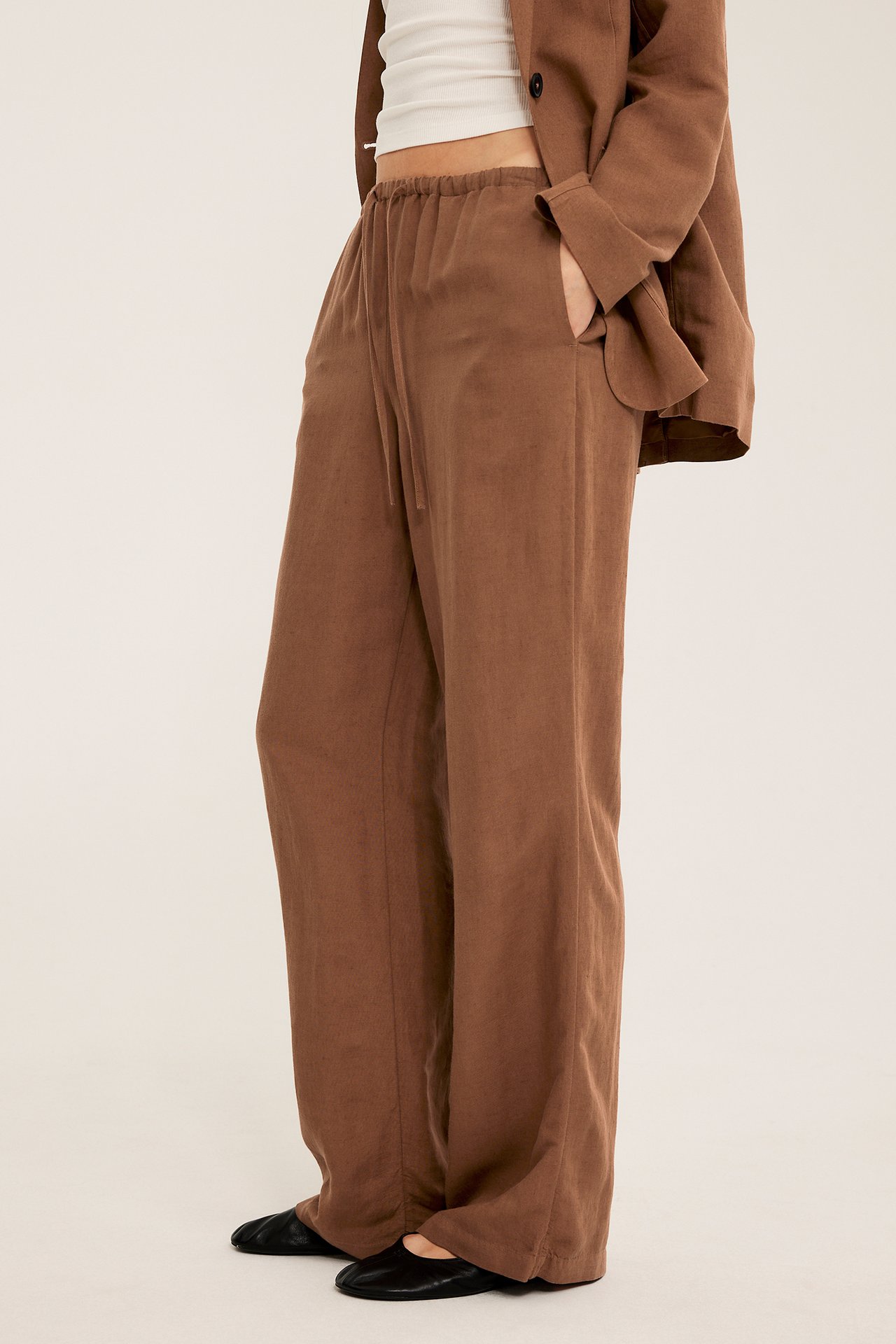 Widok z boku kobiety: brązowe lniane spodnie z troczkiem, biały top, brązowa marynarka i czarne buty.