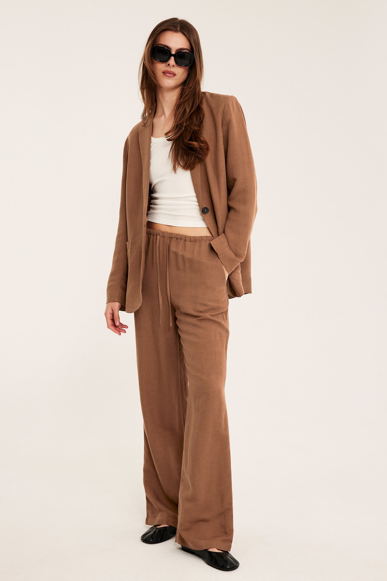 Kobieta w brązowych lnianych spodniach ze sznurkiem, pasującej marynarce, białym topie, okularach i czarnych butach.