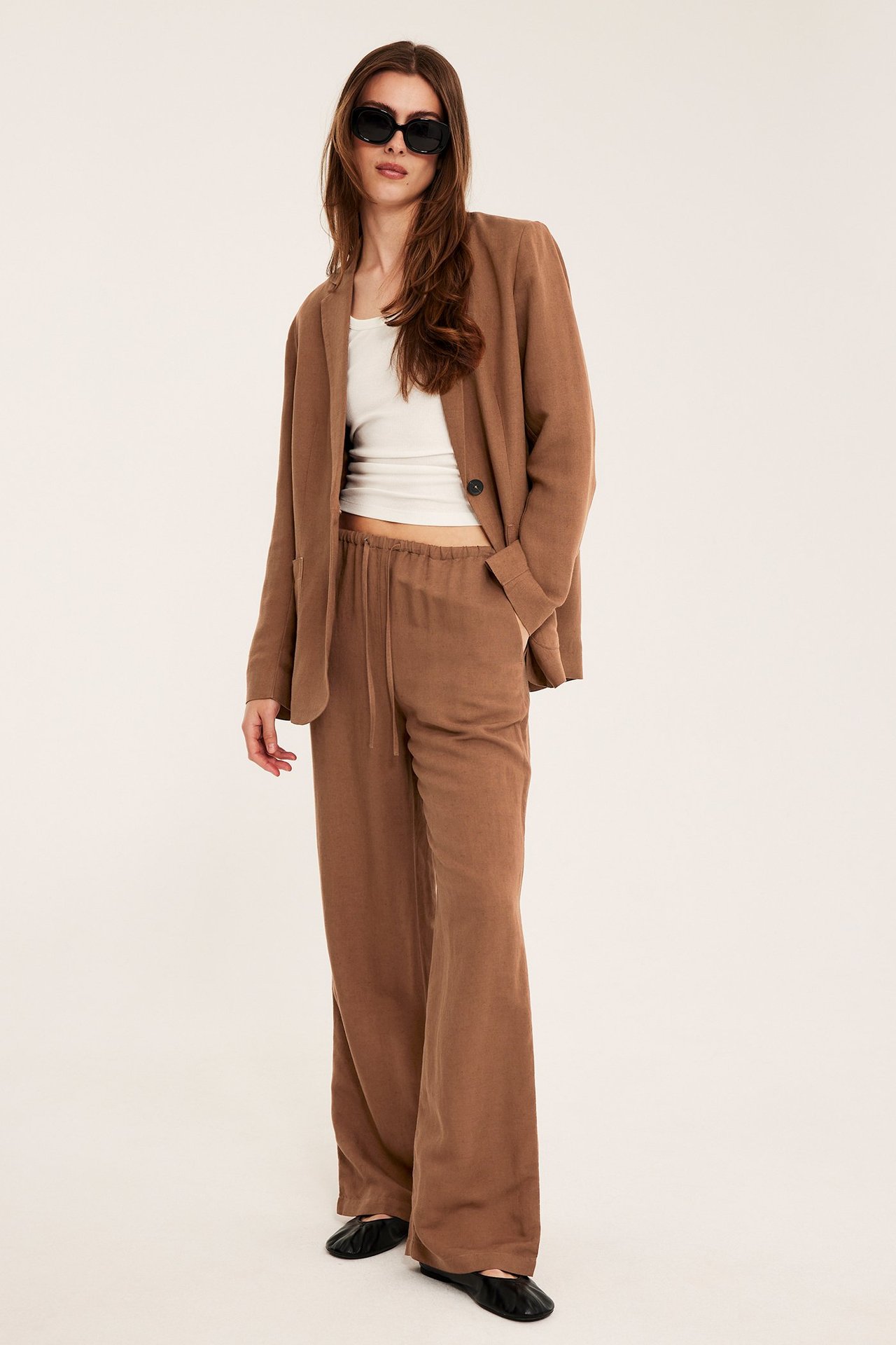 Nainen ruskeissa pellavanyörityshousuissa, yhteensopiva bleiseri, valkoinen toppi, aurinkolasit ja mustat kengät.