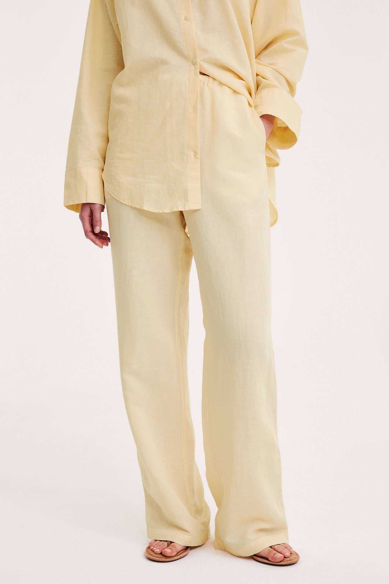 Etunäkymä: nainen yllään vaaleankeltaiset pellavasekoitehousut nyörillä, yhteensopiva paita ja ruskeat sandaalit.