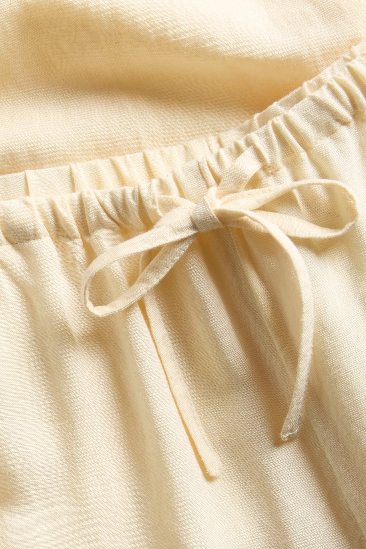 Zbliżenie na jasne, żółte spodnie z mieszanki lnu, z elastycznym pasem i sznurkiem zawiązanym w kokardę.