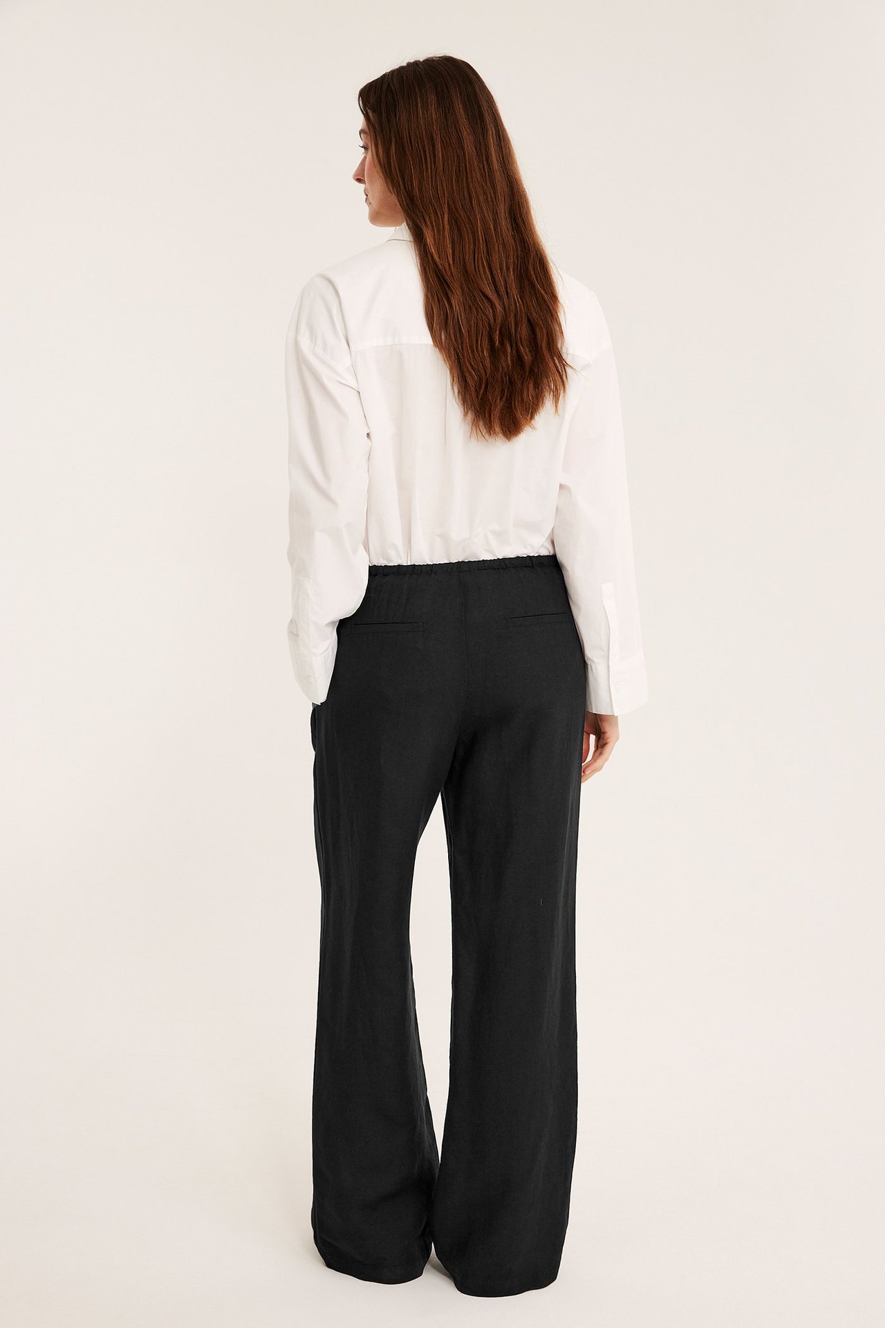 Bakfra av kvinne i svarte rette linbuksene med jukselommer og hvit skjorte.