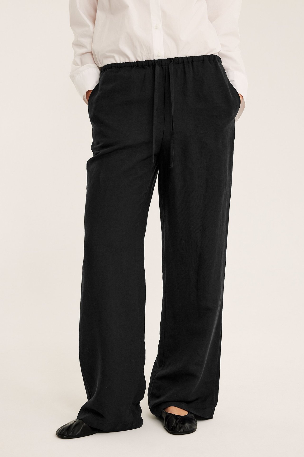 Edestä, nainen mustissa pellavasekoitehousuissa, joissa nyöri, valkoisessa paidassa ja mustissa ballerinakengissä.