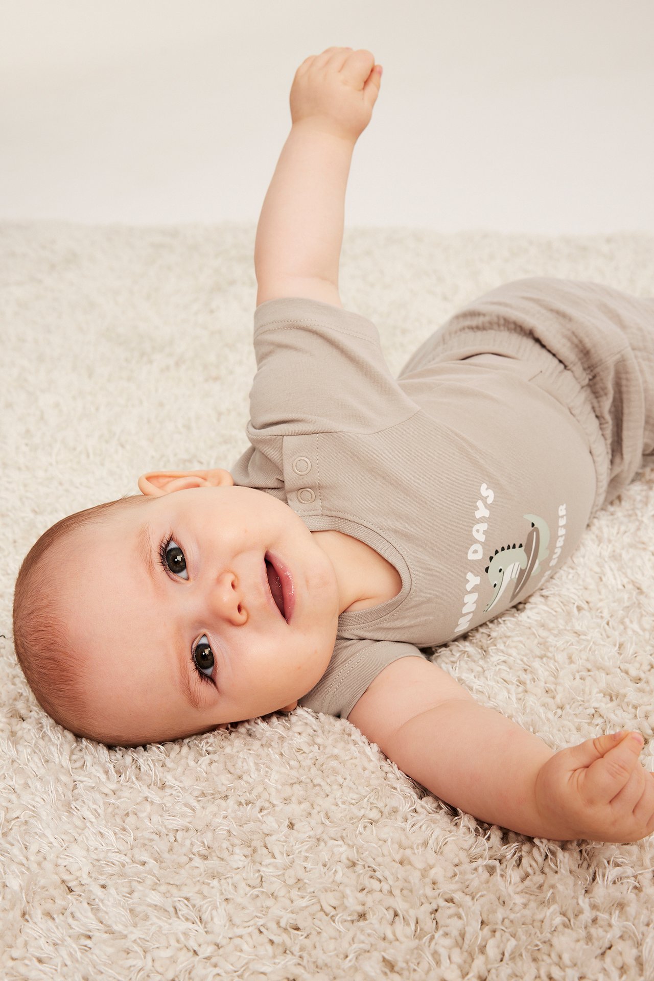 Widok z przodu: niemowlę w beżowym, krótkim body z nadrukiem krokodyla i zatrzaskami na ramieniu.