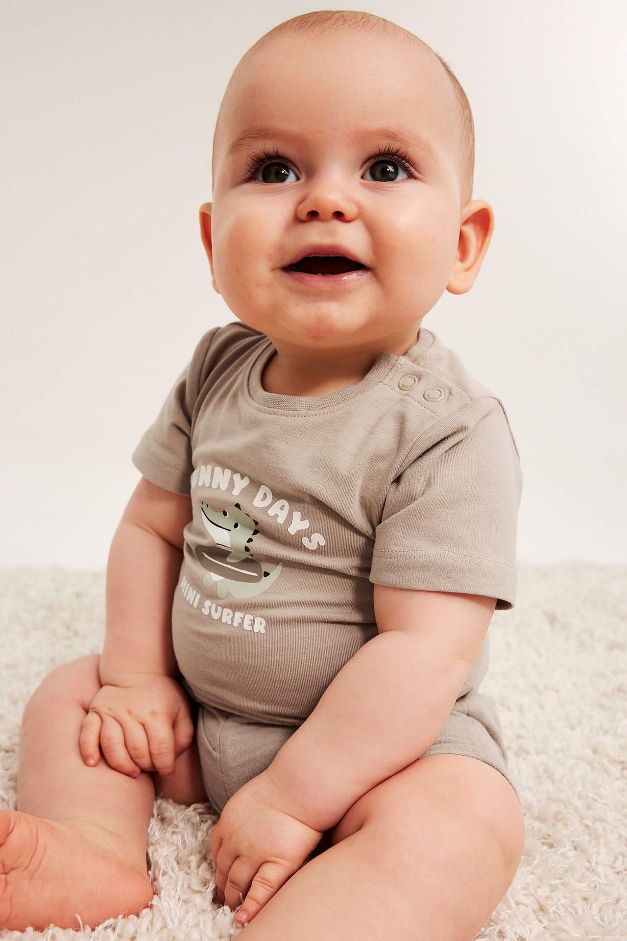 Frontbilde av en baby i en beige kortermet body med krokodilletrykk og trykknapper.