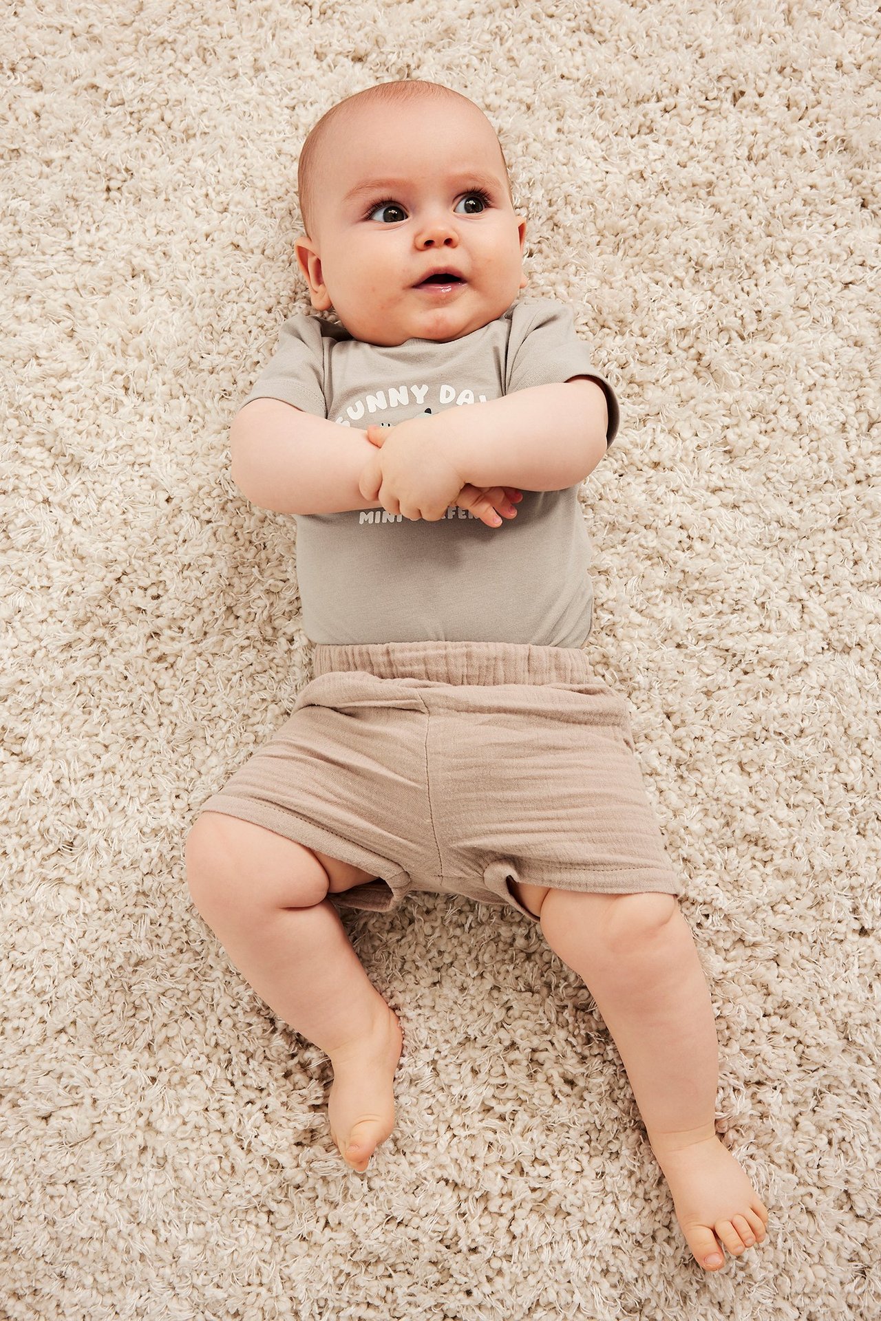 Ovenfra: Baby i en beige kortermet body med teksttrykk, trykknapper og ensfargede beige shorts.