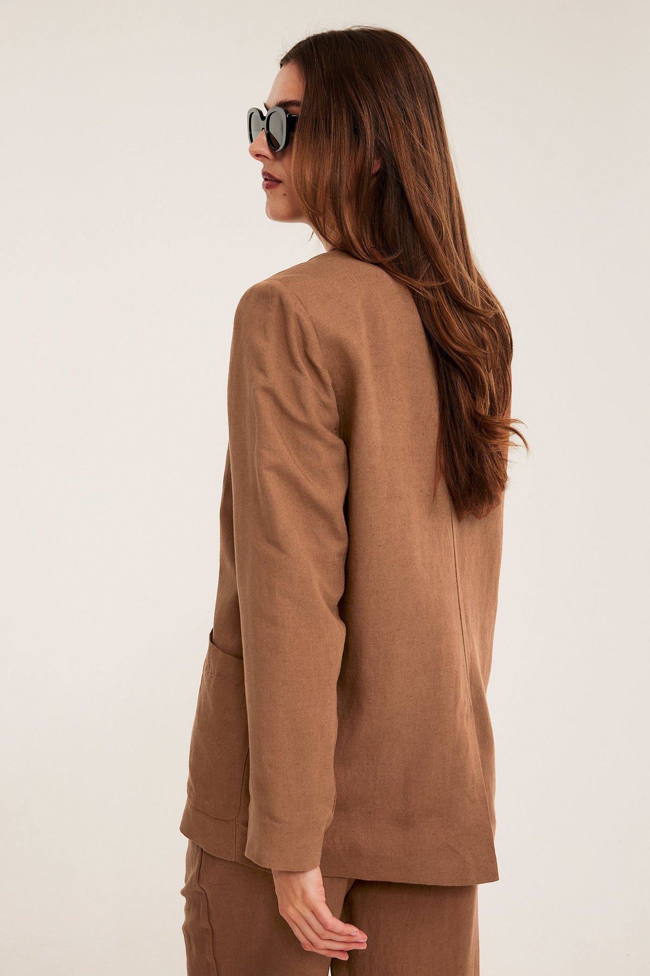 Nainen ruskeassa löysässä pellavasekoitekavajassa paikkataskuilla, yhteensopivissa housuissa ja mustissa aurinkolaseissa.