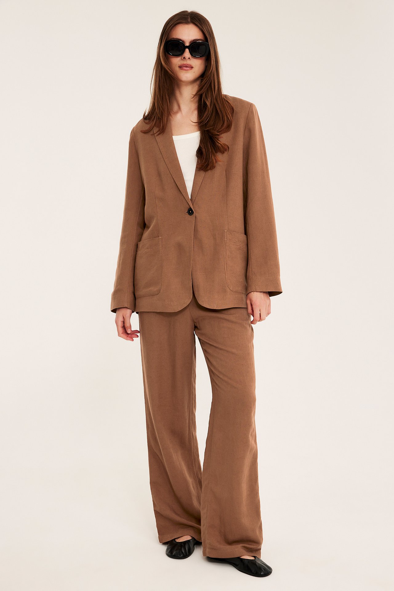 Nainen yllään löysä ruskea pellavablazer, yhteensopivat housut, valkoinen toppi ja aurinkolasit.