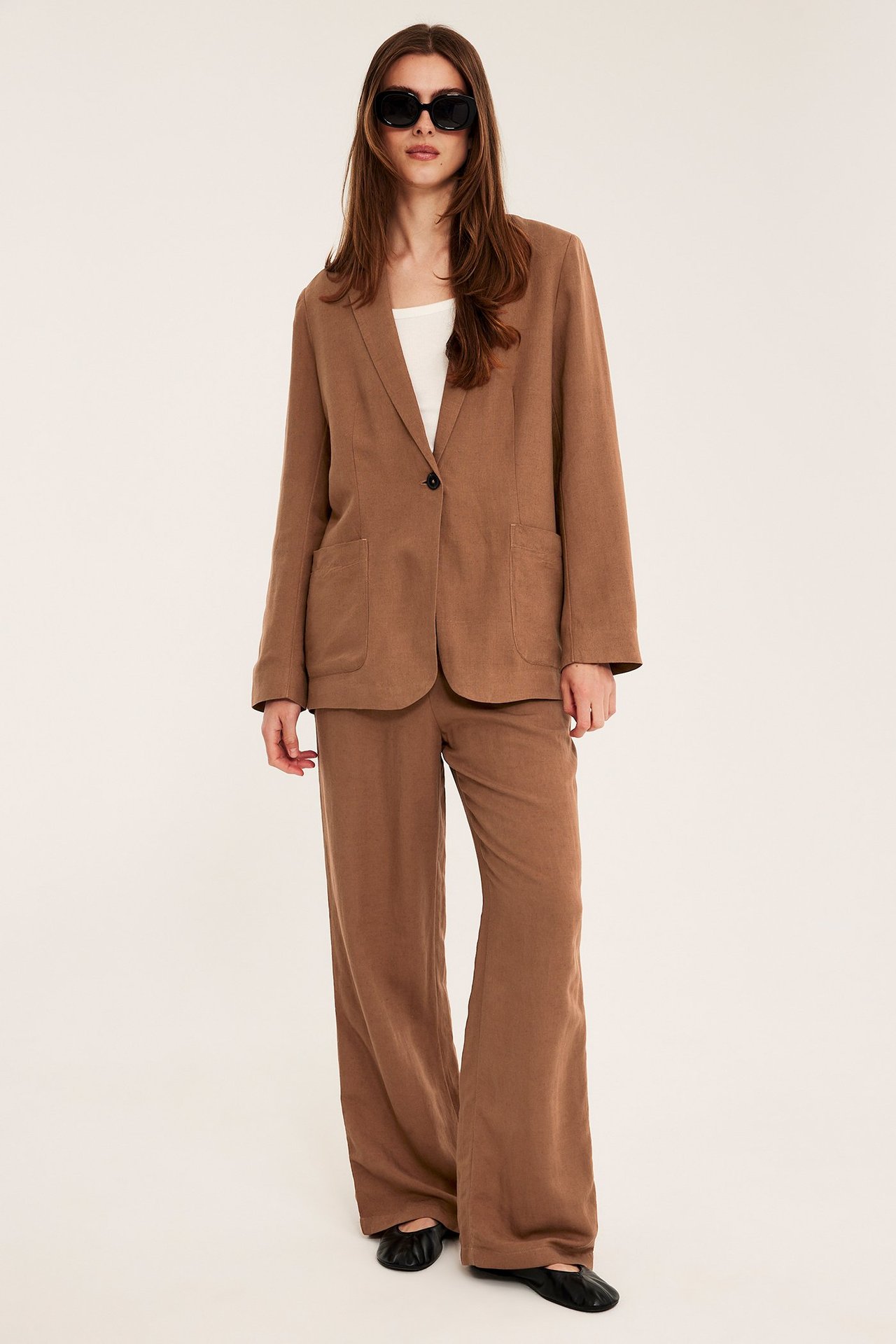 Nainen yllään löysä ruskea pellavablazer, yhteensopivat housut, valkoinen toppi ja aurinkolasit.