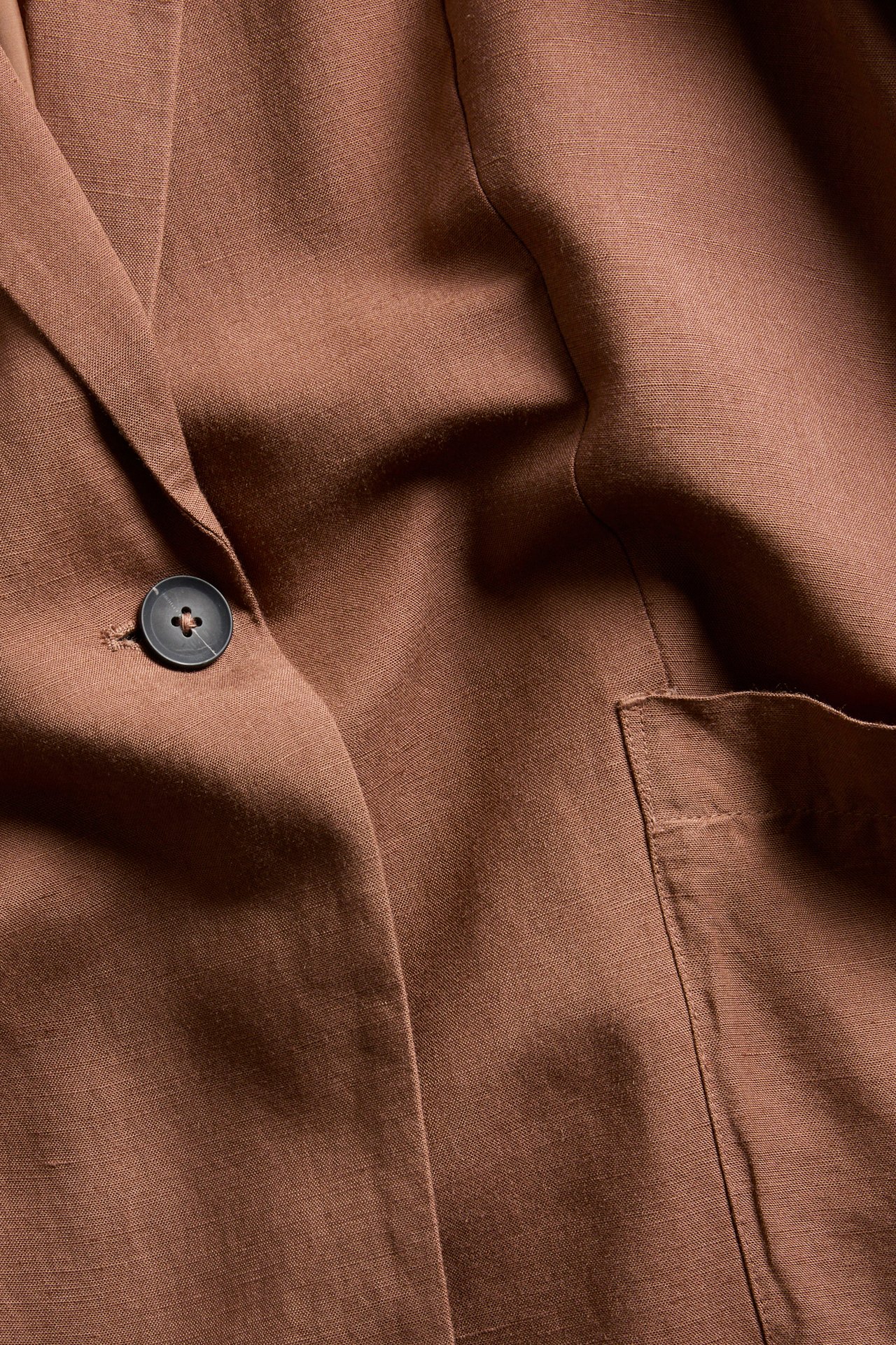 Närbild av en brun kavaj i linnemix med avslappnad passform, en mörk knapp och en utanpåliggande ficka.