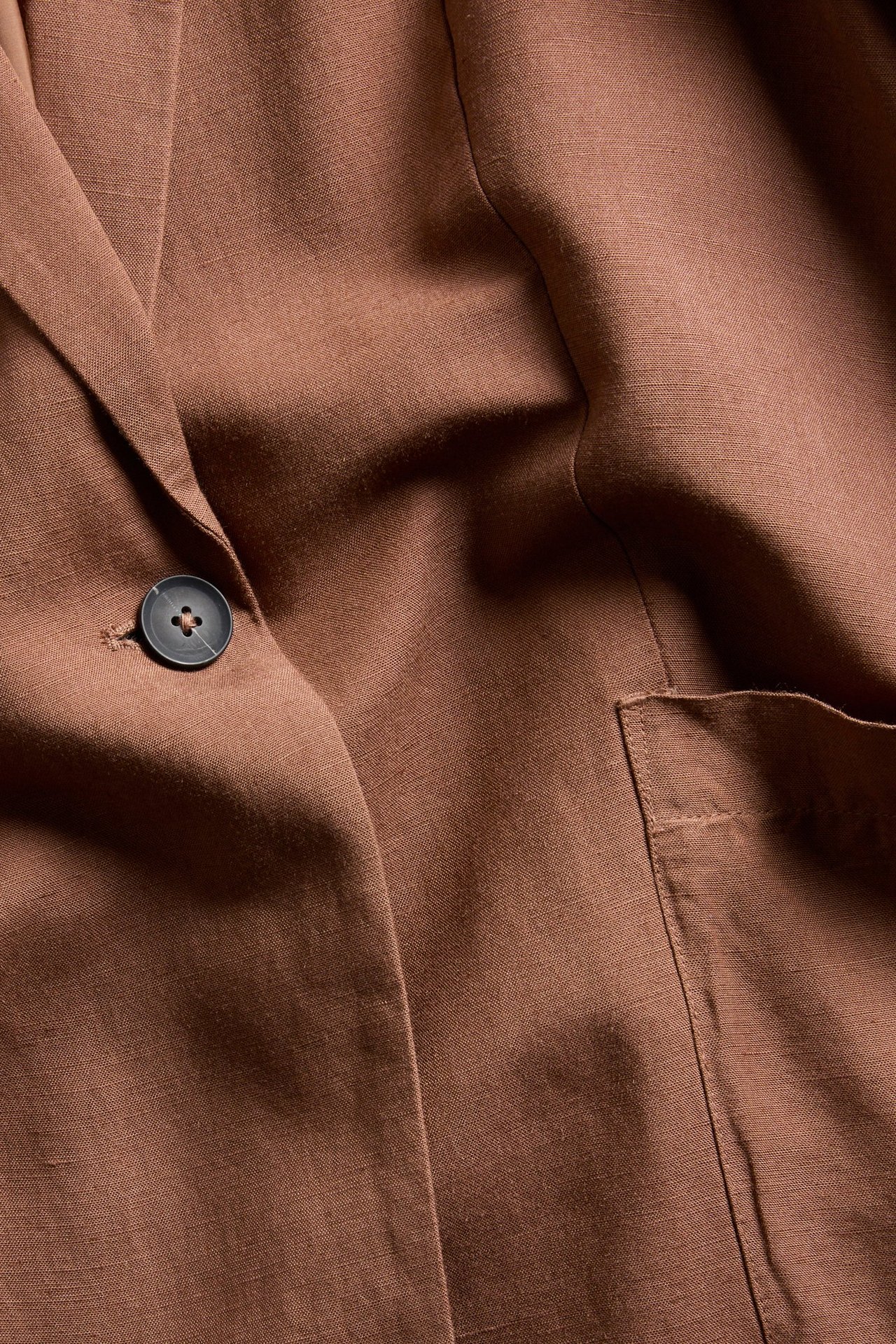 Zbliżenie na brązową marynarkę z mieszanki lnu, o luźnym kroju, z jednym ciemnym guzikiem i naszywaną kieszenią.