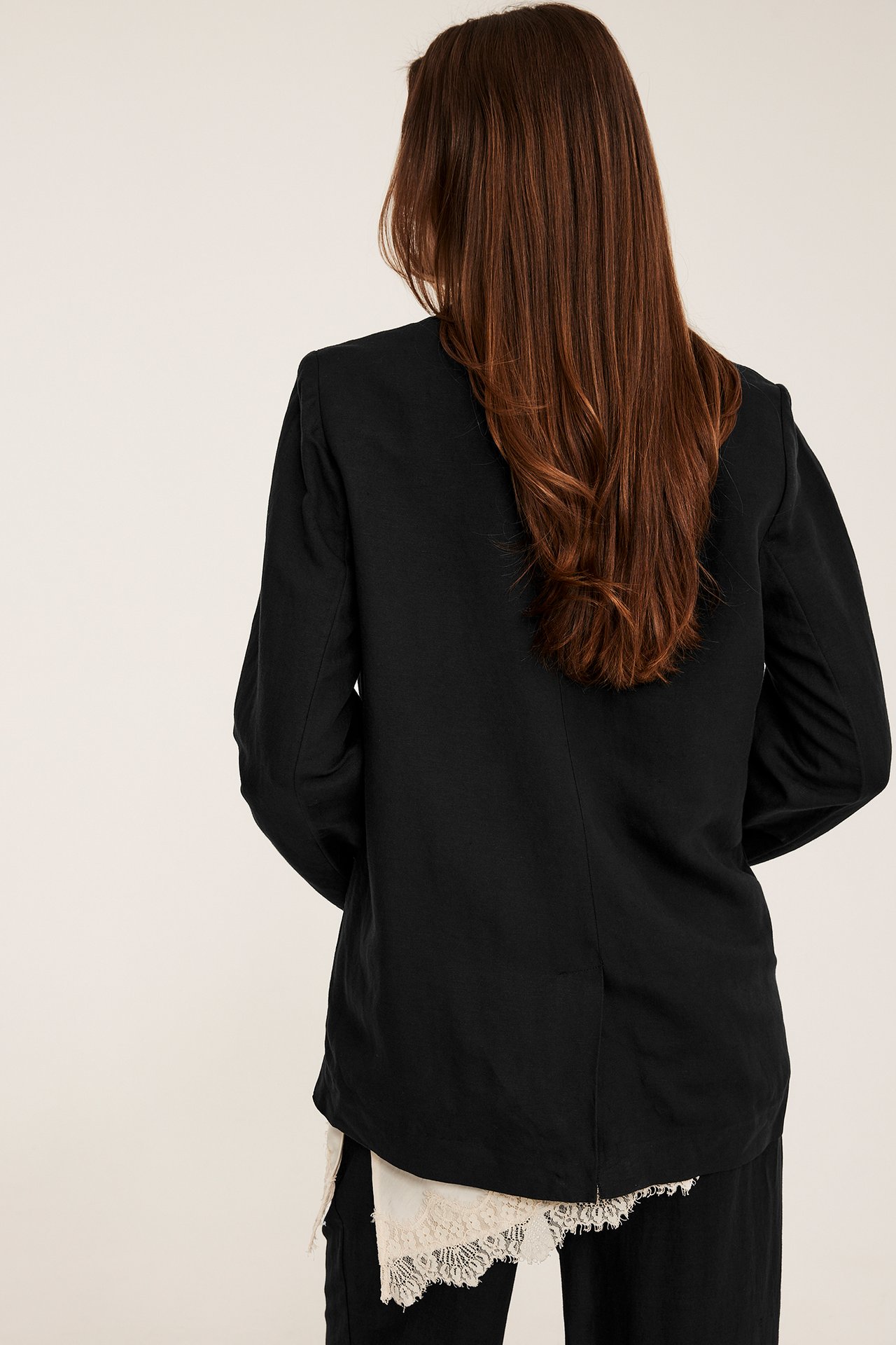 Nainen takaapäin mustassa, rento mallisessa pellavasekoitekavajassa, jonka alla vaalea pitsipaitapusero ja mustat housut.