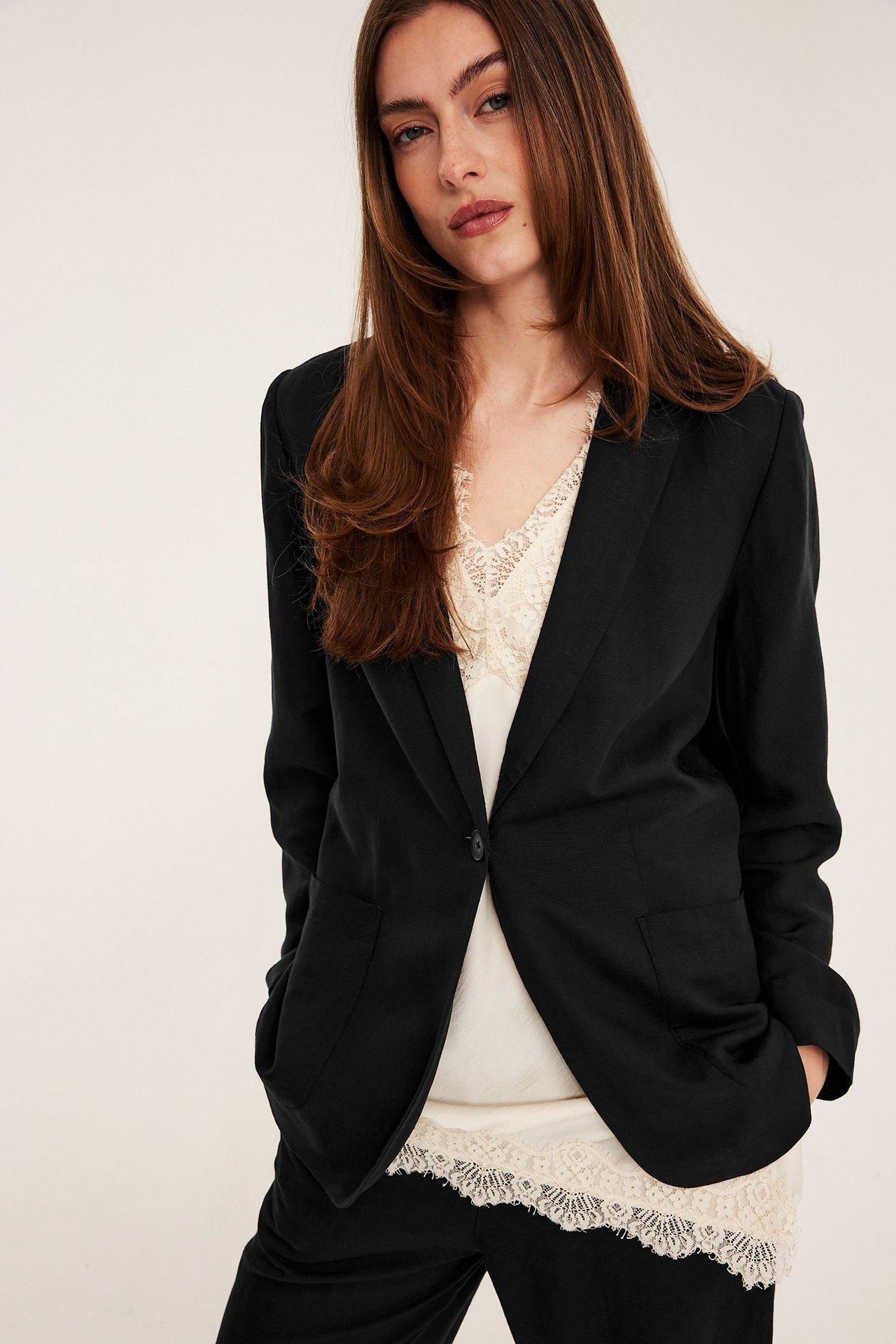 Edestä: Nainen mustassa pellavasekoitekavajassa yhdellä napilla, päällä kermanvärinen pitsitoppi ja mustat housut.