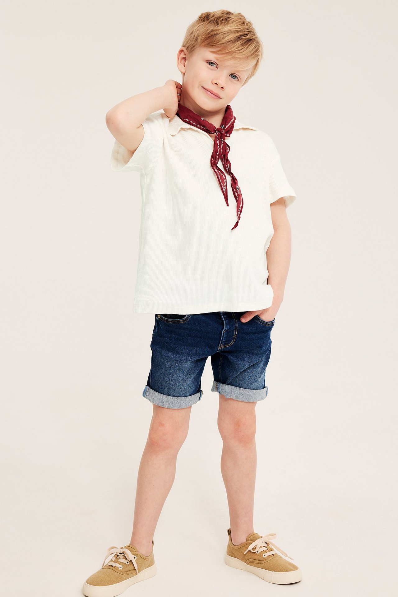 Etunäkymä lapsesta, jolla on luonnonvalkoinen strukturoitu puuvillapolo, punainen huivi, siniset farkkushortsit ja beiget tennarit.