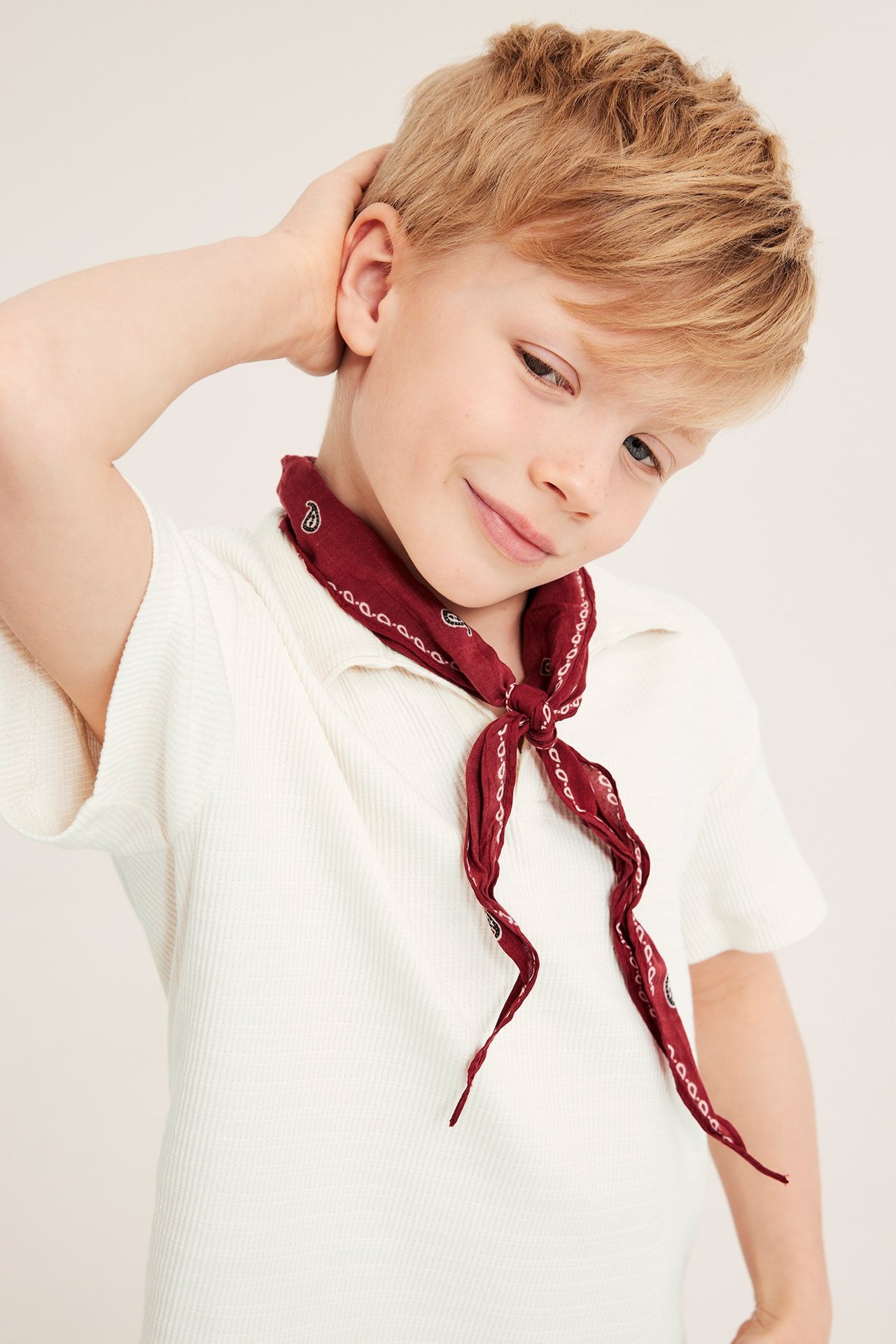 Nærbilde av et smilende barn med en offwhite ribbet bomulls-pologenser og en rød paisley-bandana.
