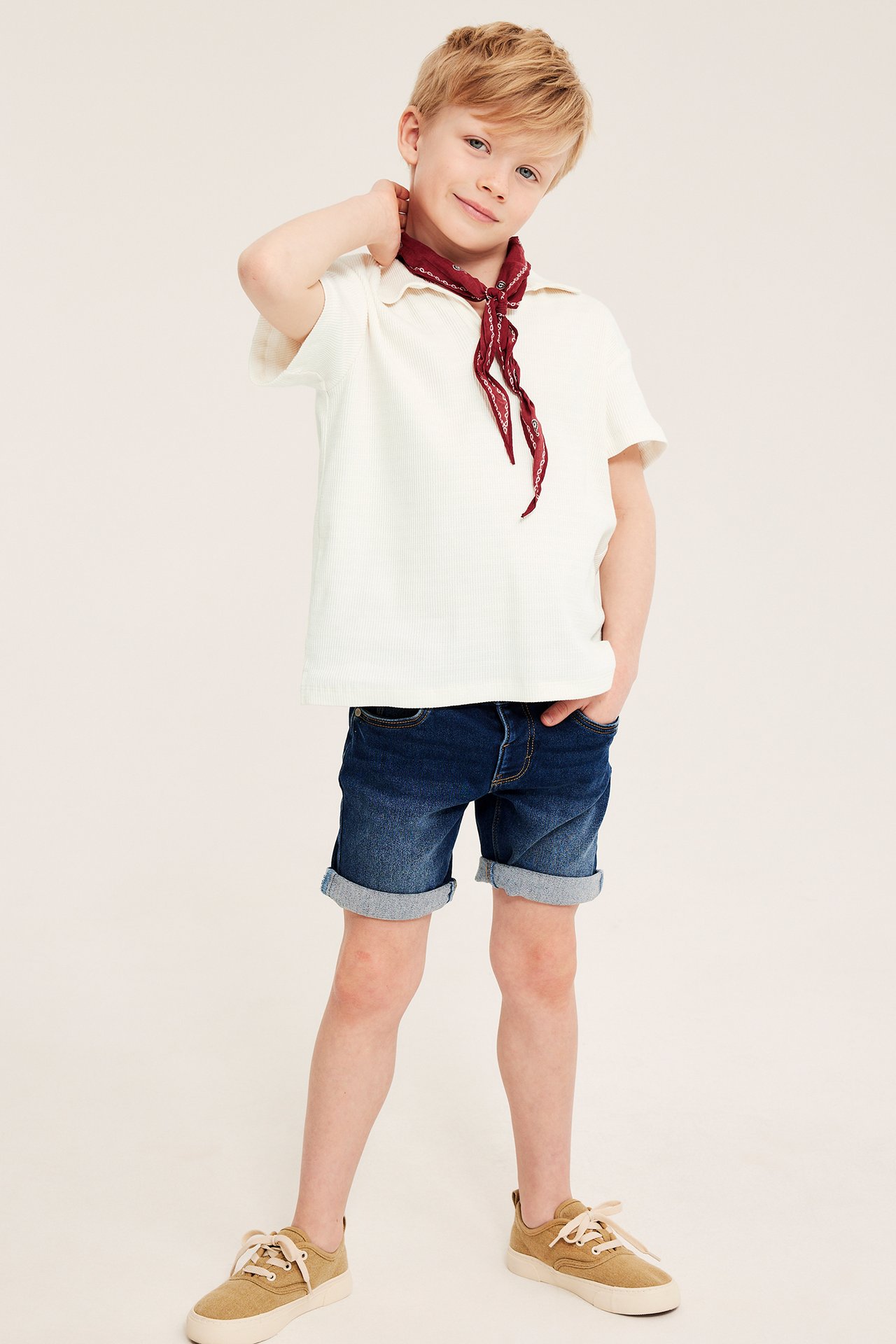 Frontbilde av et barn i offwhite strukturert bomullspolo, rødt skjerf, blå dongerishorts og beige sneakers.