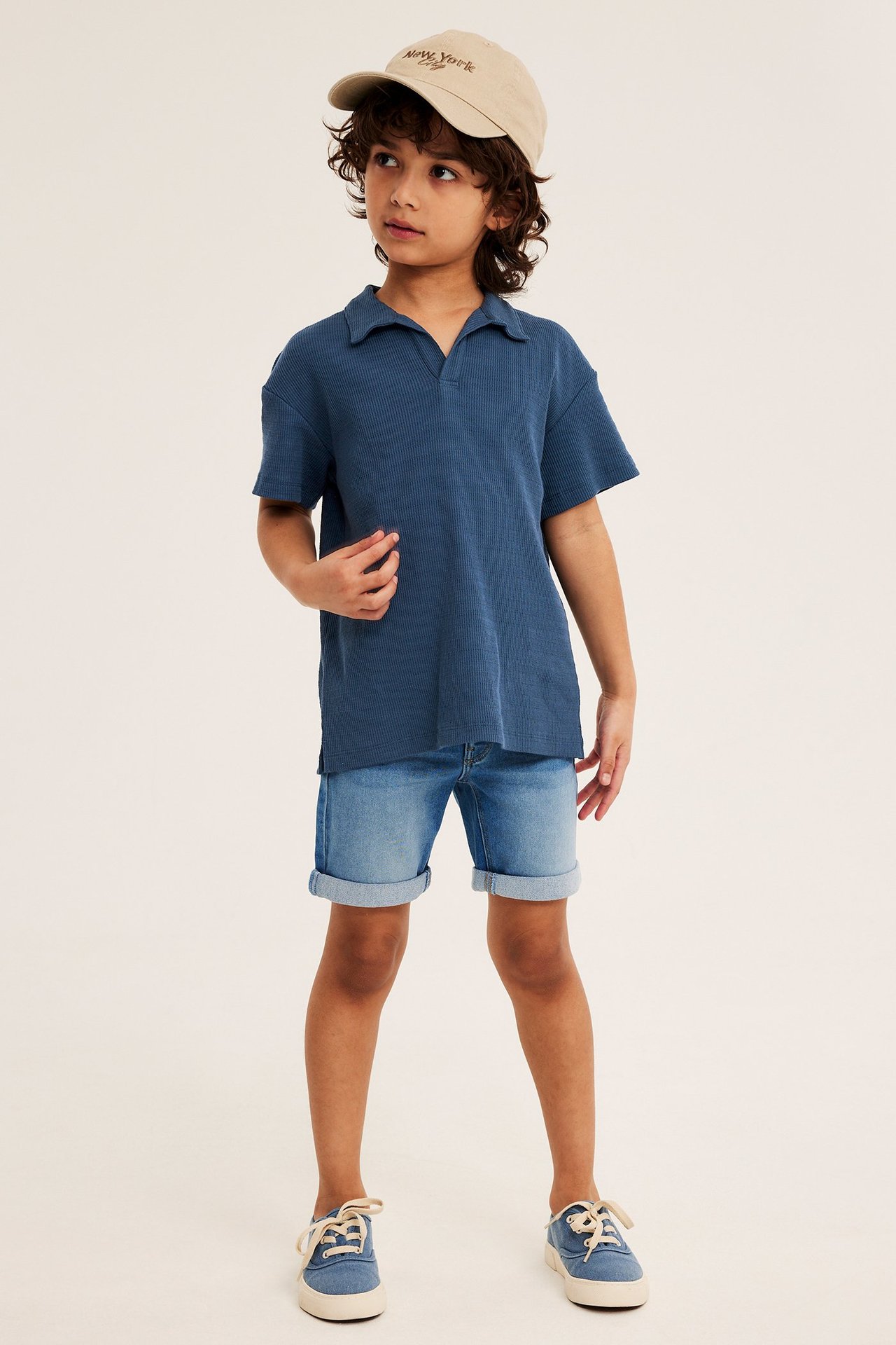 Ett barn bär en blå strukturstickad pikétröja, ljusblå denimshorts, blå sneakers och en beige keps.