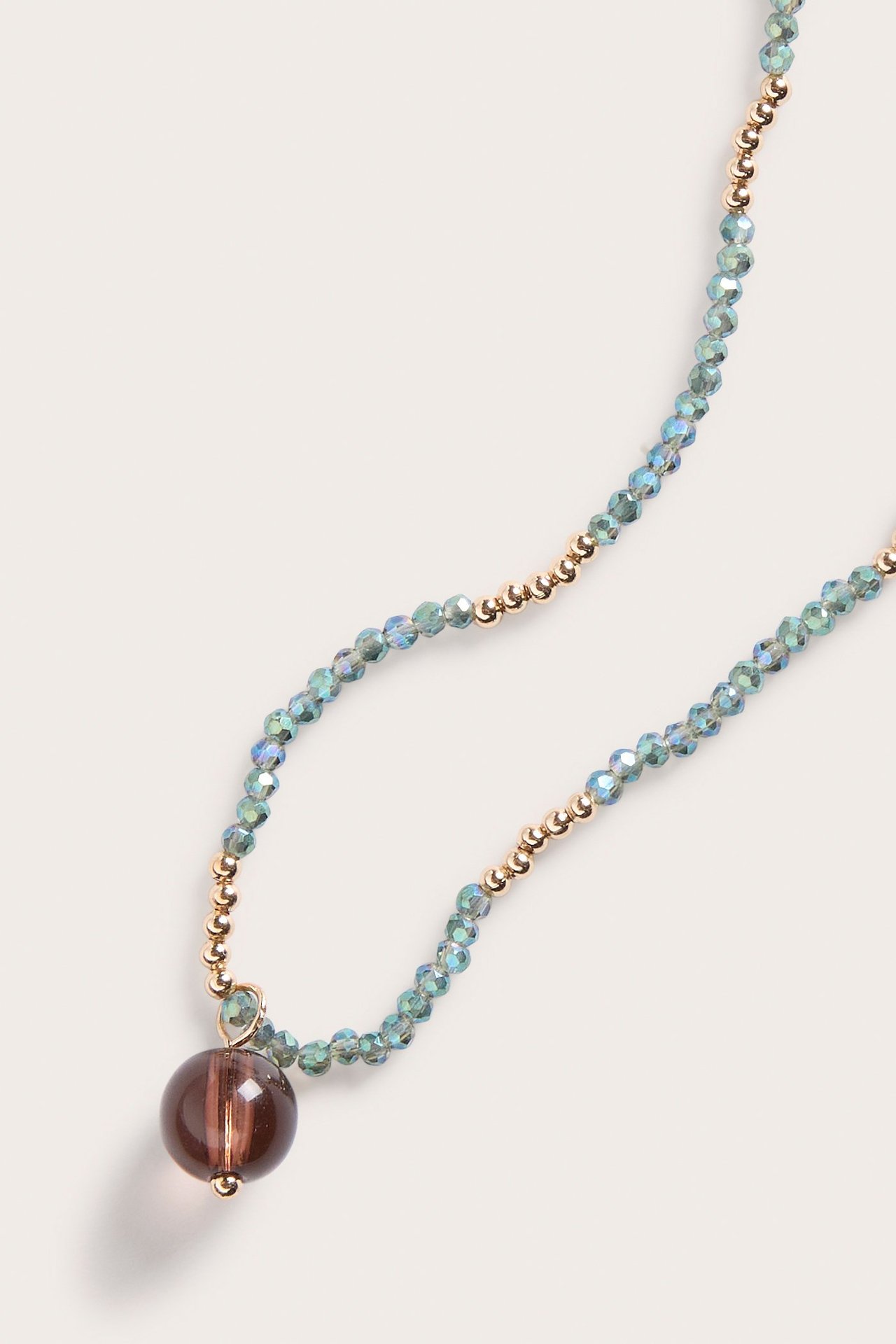Zbliżenie naszyjnika z opalizującymi zielono-niebieskimi szklanymi koralikami, małymi złotymi koralikami i dużym brązowym szklanym wisiorkiem.