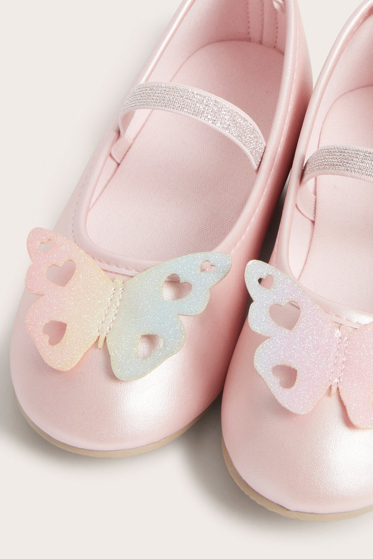 Zbliżenie na jasnoróżowe metaliczne balerinki dziecięce z opalizującymi brokatowymi motylami i srebrnymi elastycznymi paskami.