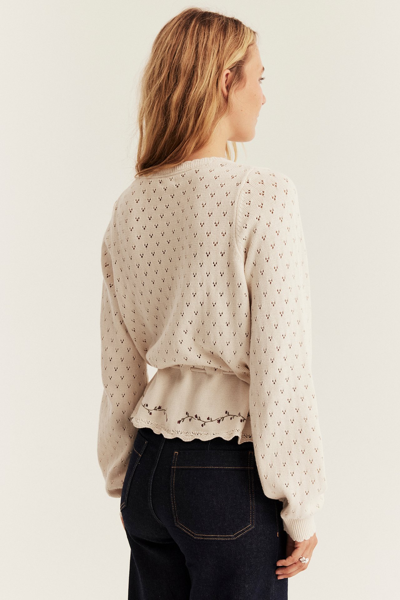 Ryggvy av kvinnlig modell i beigefärgad ögonstickad tröja med broderad peplum över mörkblå jeans.
