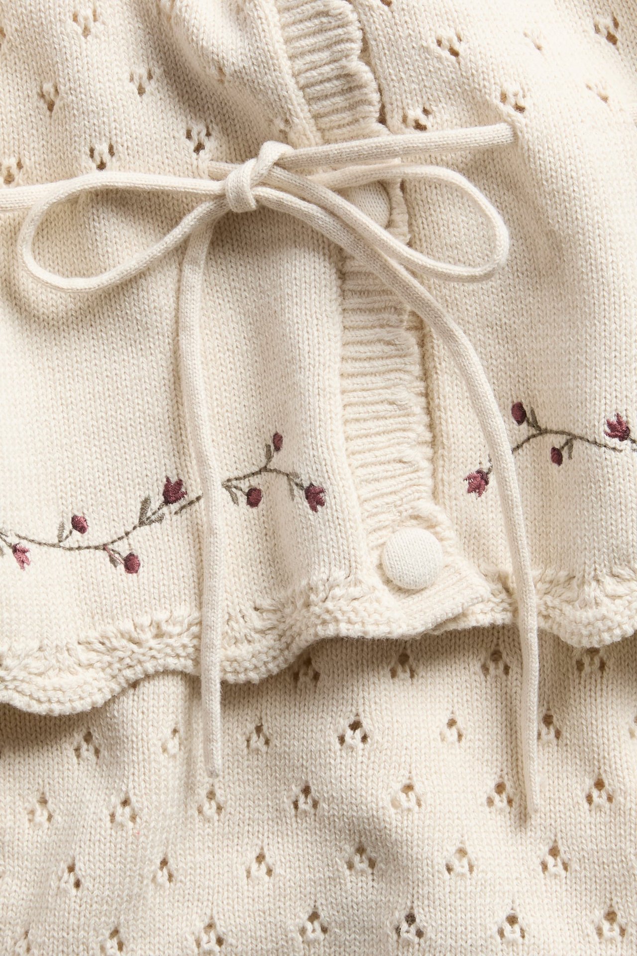 Närbild på gräddvit stickad kofta med blommigt broderi och rosett över hålmönstrad tröja.