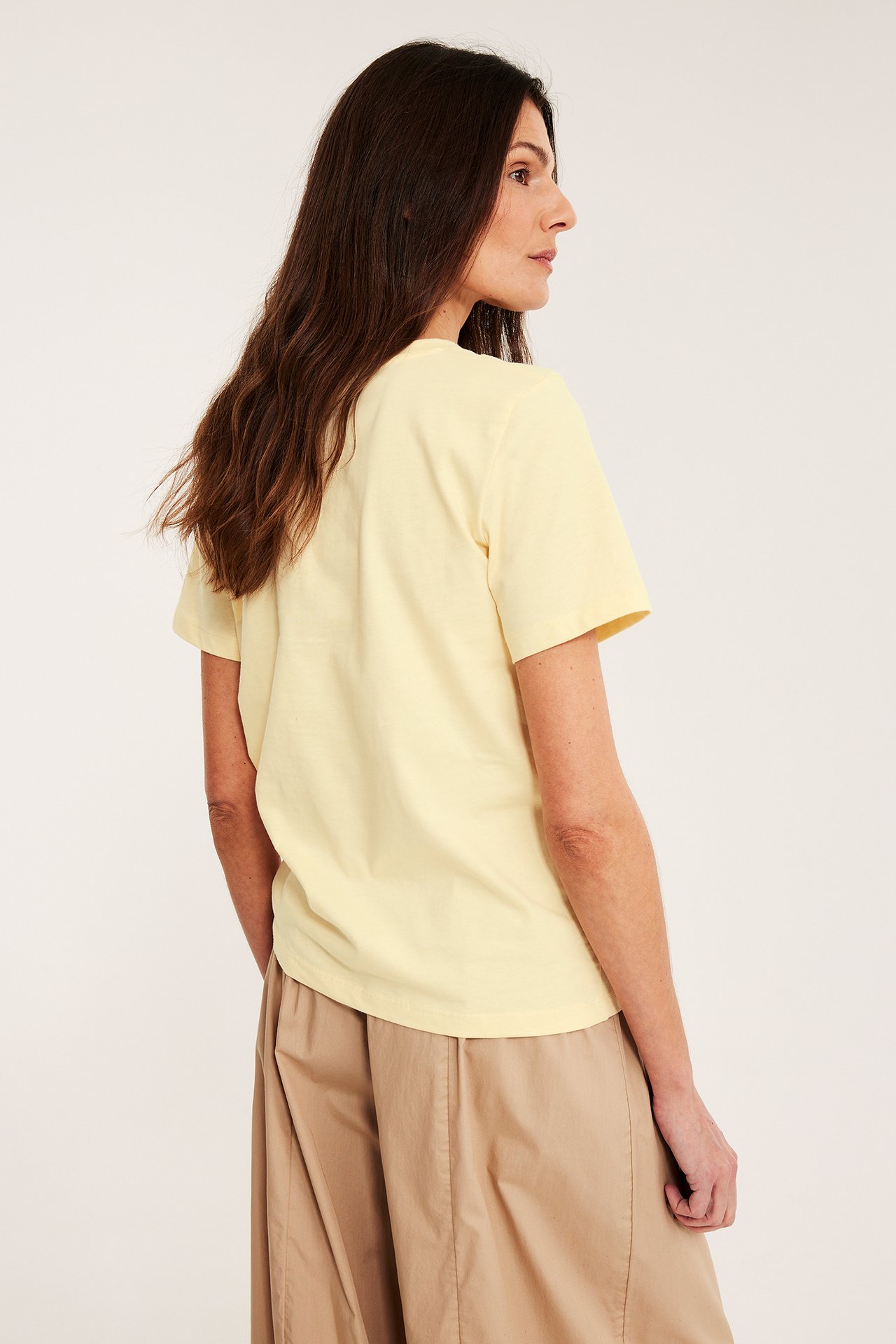 Sida-bakifrånvy av en kvinna i ljusgul, rak bomulls-T-shirt och ljusbruna vida byxor.