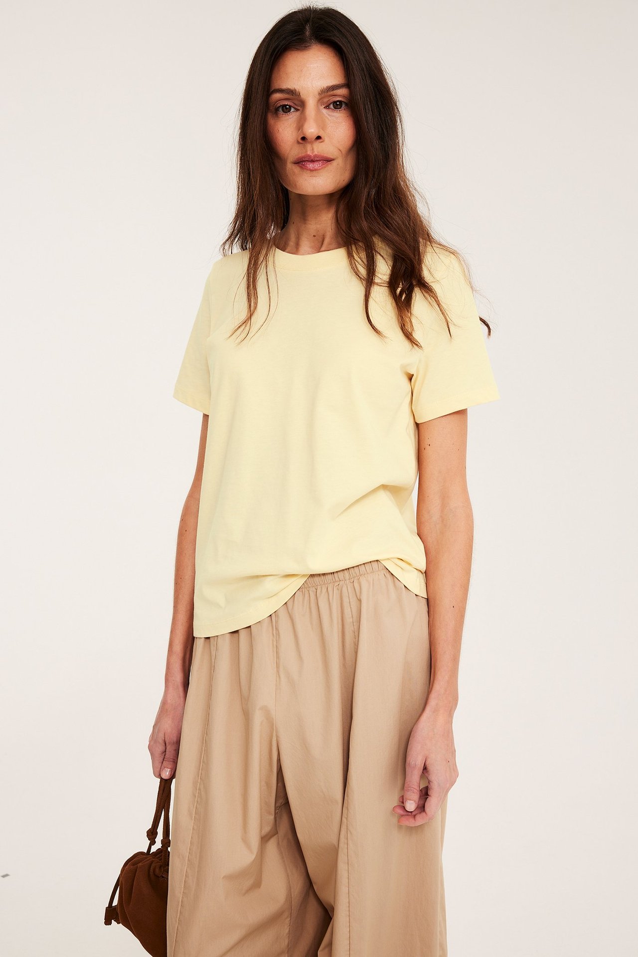 Forfra: Kvinne i lys gul, rett passform bomulls-T-skjorte, beige bukser, holder en brun veske.