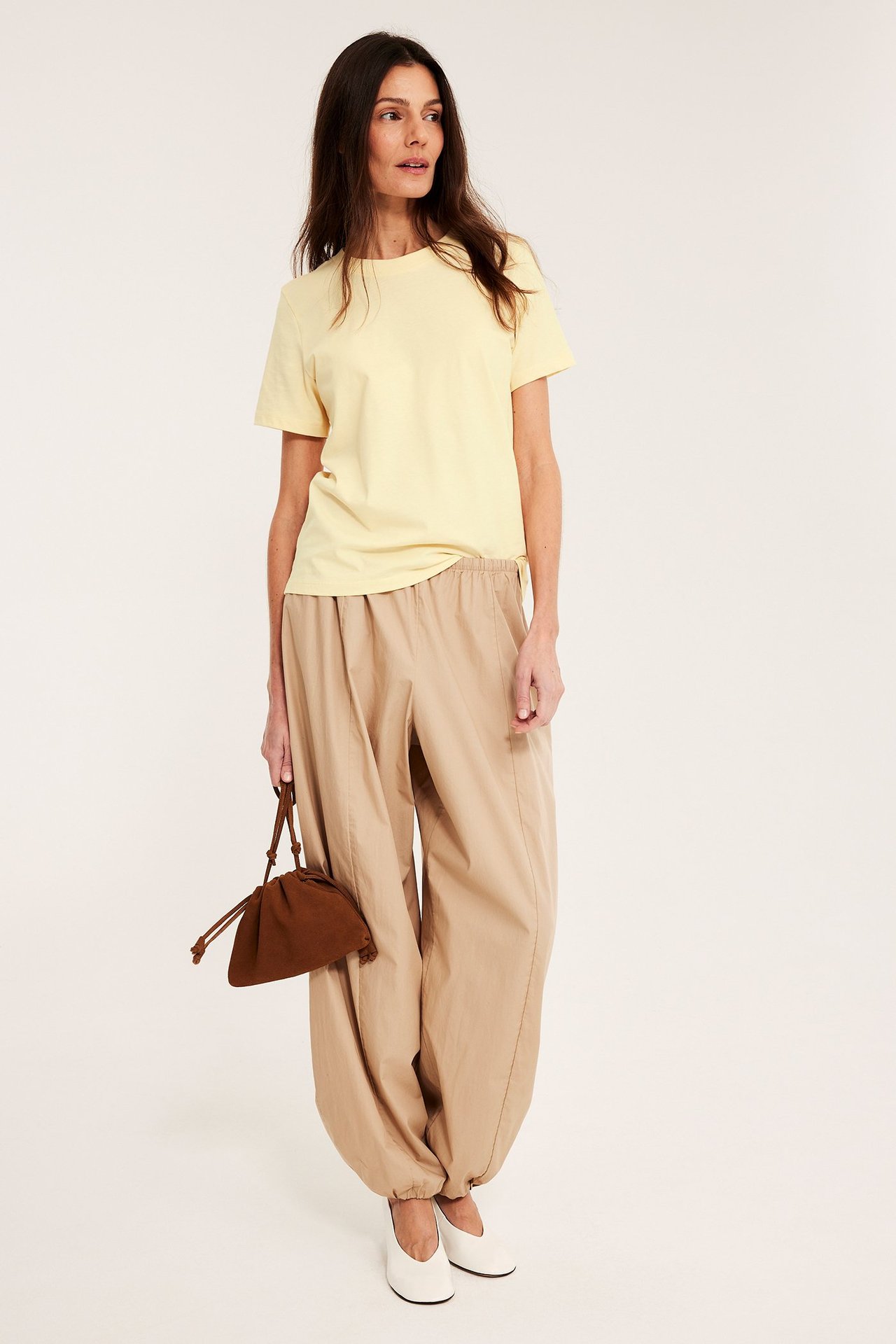 Frontvisning: Kvinne i lys gul T-skjorte, beige ballongbukser, hvite flate sko, holder brun poseveske.
