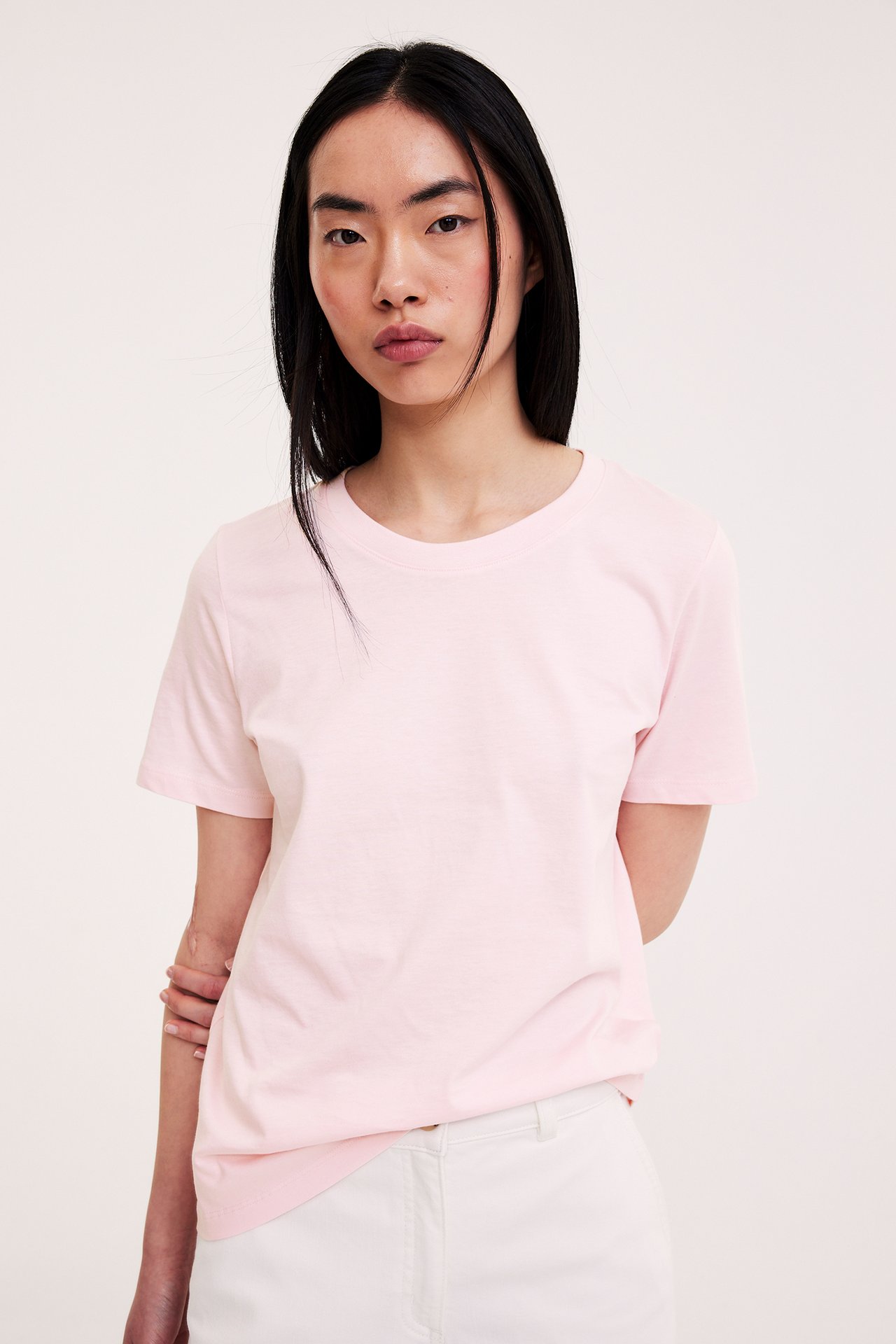 Frontbilde av en kvinne i en lys rosa, ensfarget bomulls-T-skjorte med rett passform og hvite bukser.