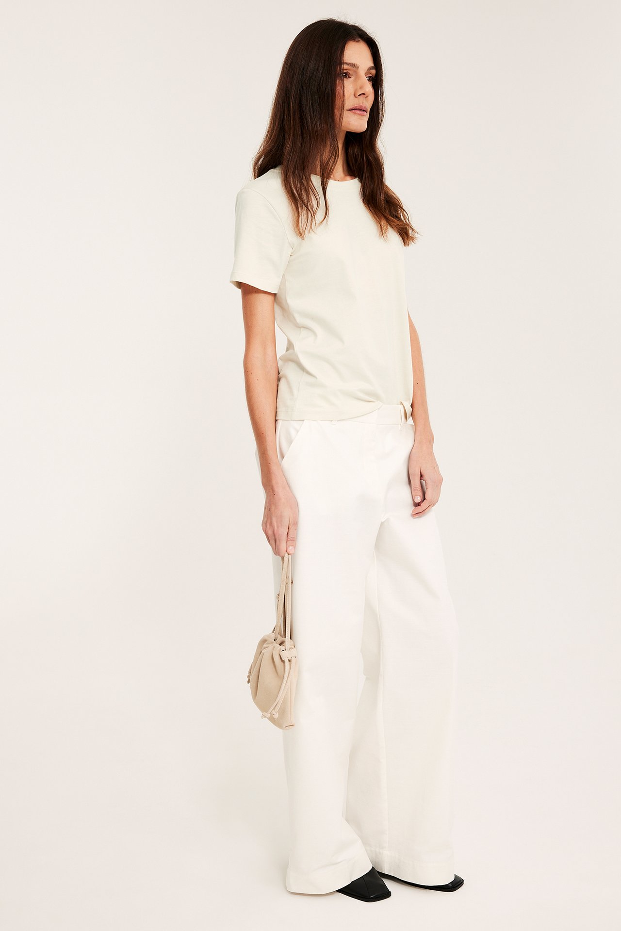 Naisen sivukuva yllään luonnonvalkoinen puuvillainen t-paita, valkoiset leveät housut, mustat kengät, pitäen beigeä laukkua.