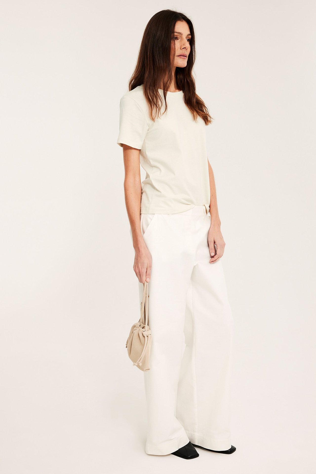 Sidebilde av en kvinne i en offwhite bomulls-T-skjorte, hvite vide bukser, svarte sko, holdende en beige veske.