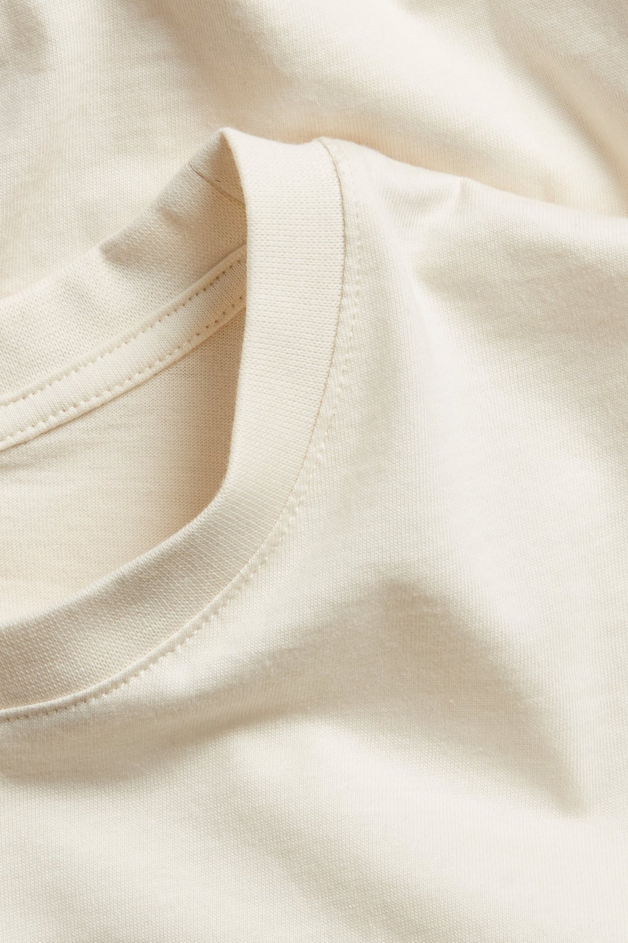 Zbliżenie na kremowy, regularny T-shirt bawełniany z prążkowanym okrągłym dekoltem i gładką fakturą materiału.