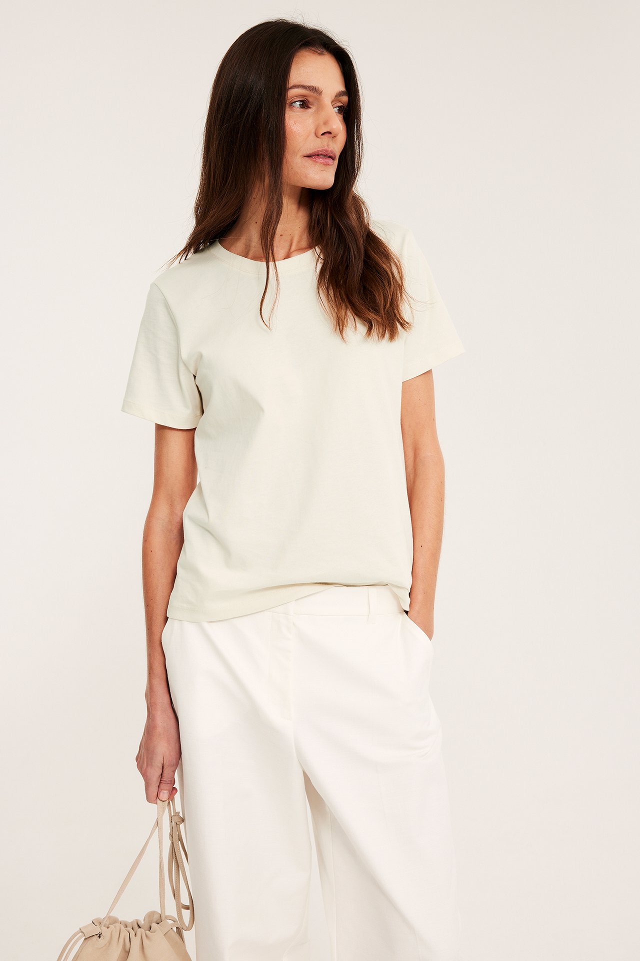 Edestäpäin nainen yllään luonnonvalkoinen suora puuvilla T-paita, valkoiset housut ja pitelee beigeä laukkua.