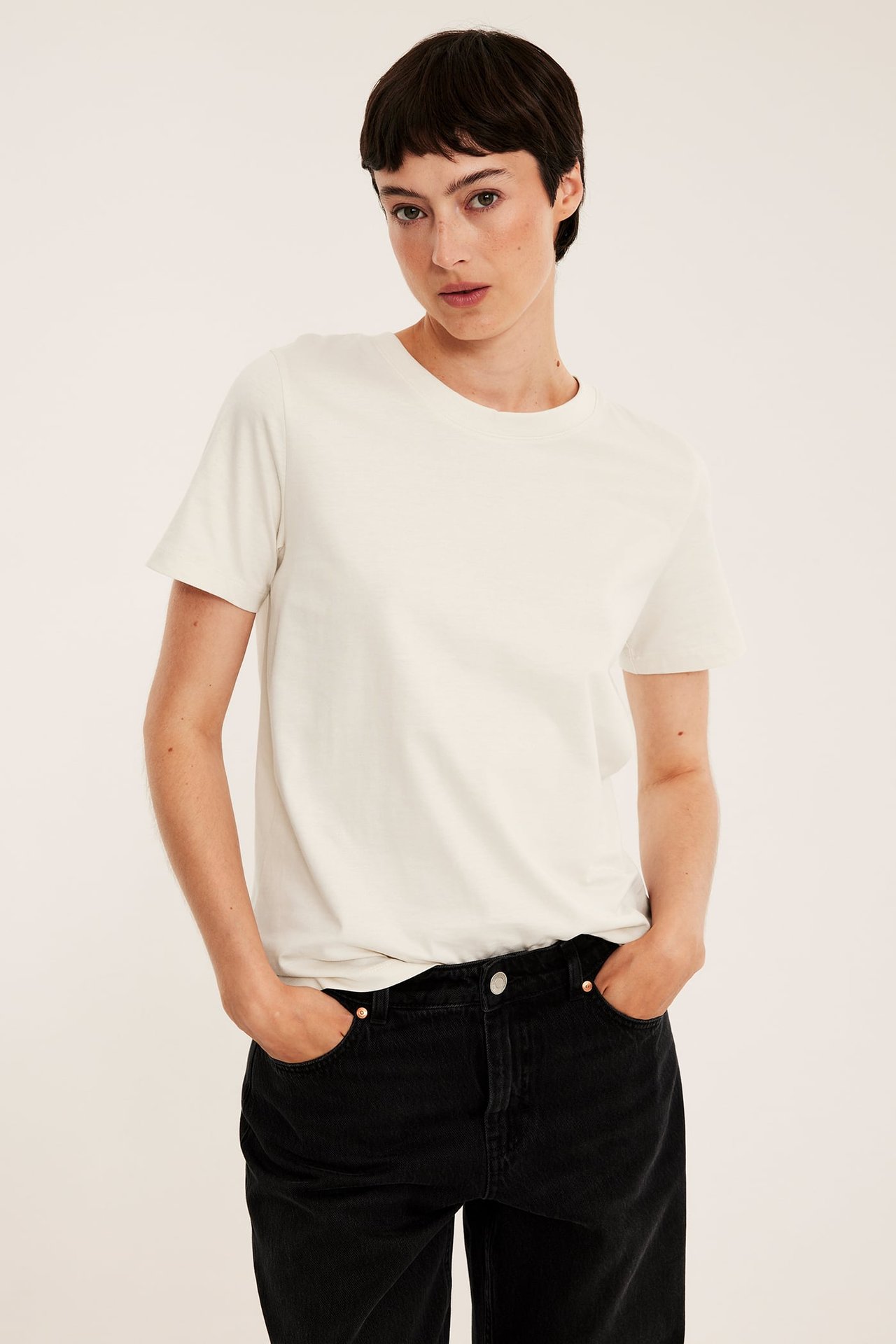 Frontvy av en kvinna i en offwhite rak bomulls-T-shirt och svarta jeans med händerna i fickorna.