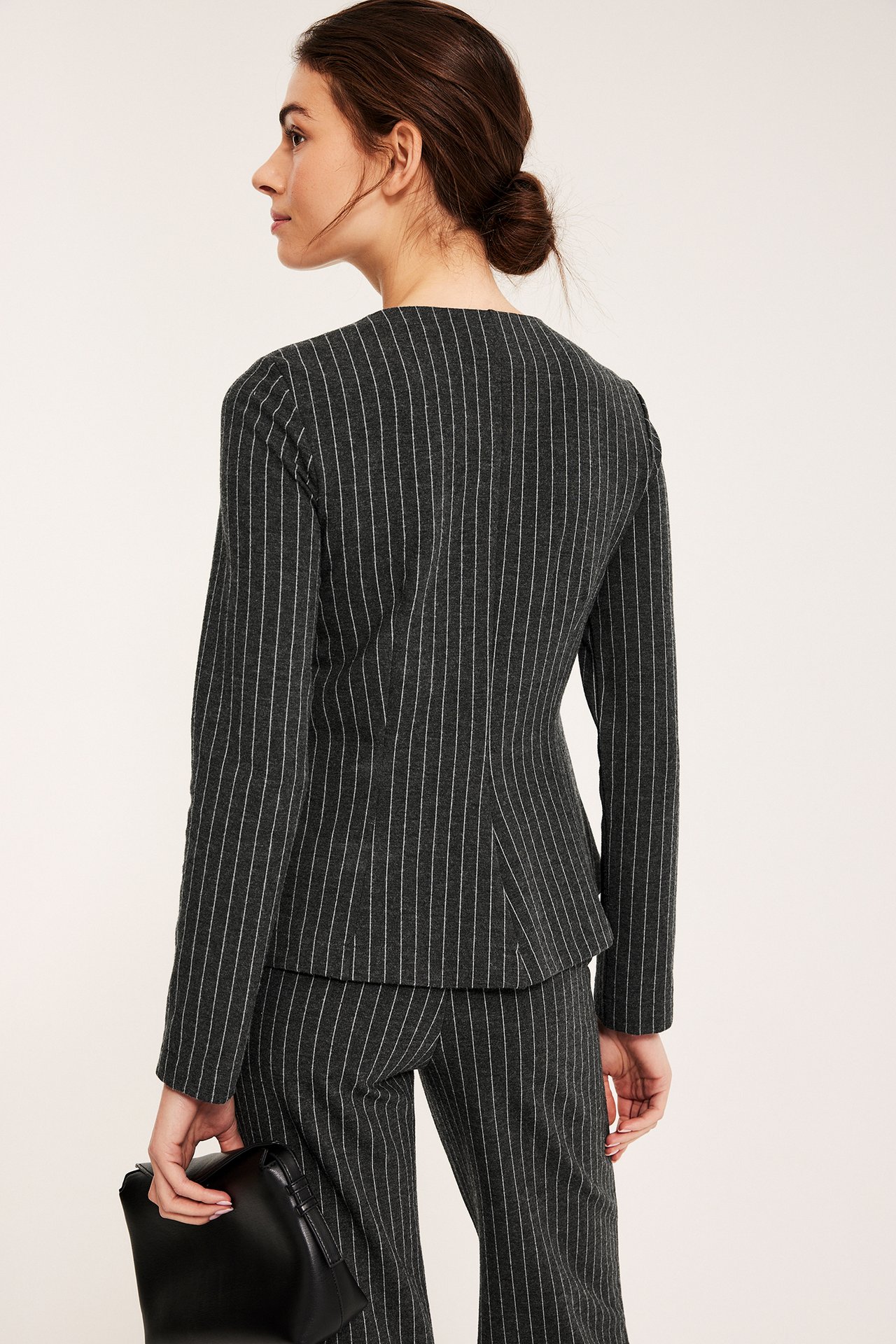 Sida vy av kvinna i mörkgrå kritstrecksrandig jerseykavaj med peplum, matchande byxor, håller svart handväska.