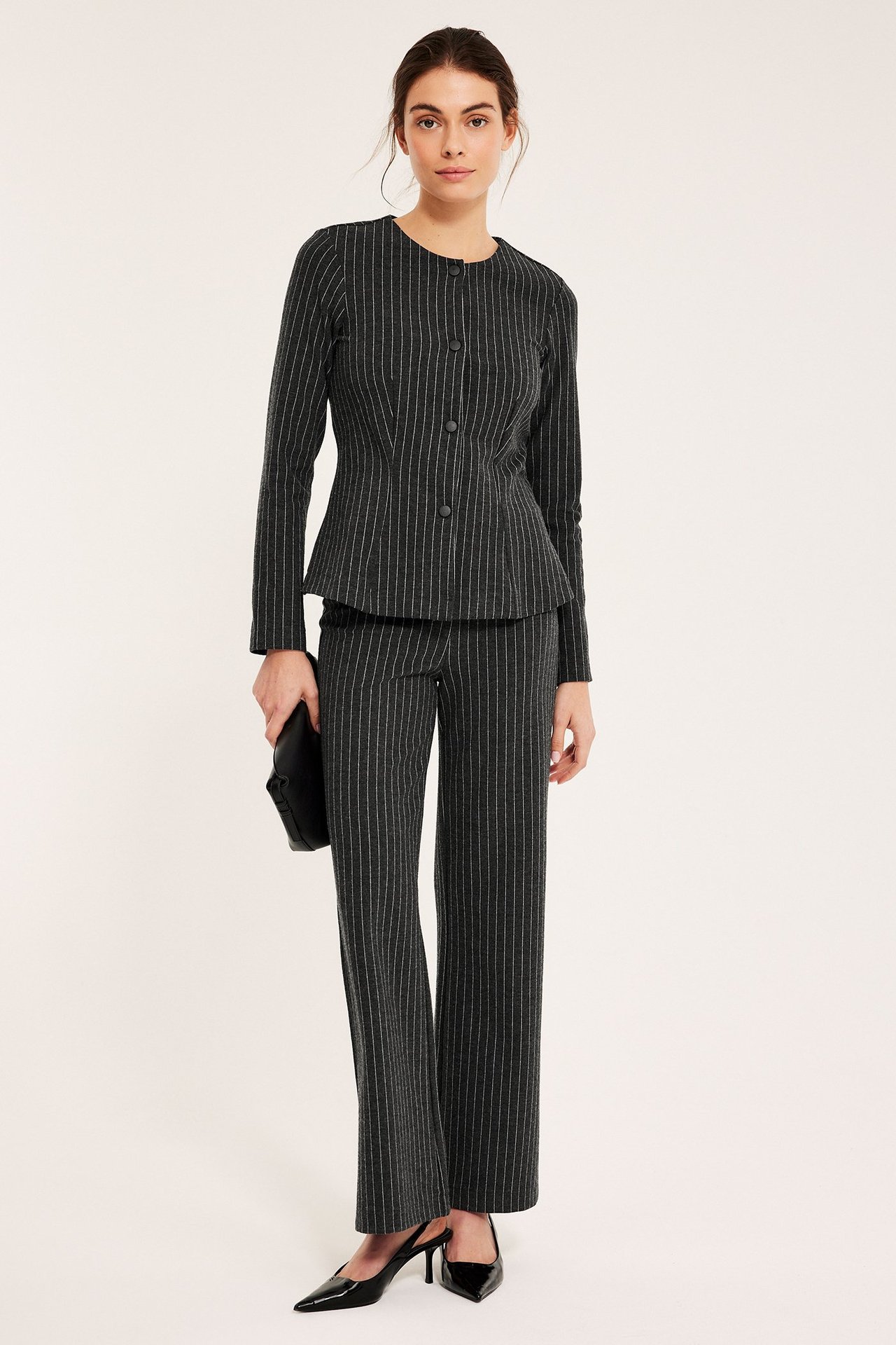 Framifrån, kvinna i mörkgrå kritstrecksrandig jerseykavaj med peplum, matchande byxor och svart clutch.