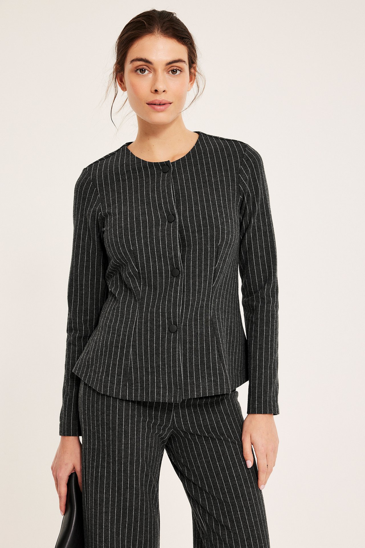 Frontvy, kvinna i mörkgrå kritstrecksrandig kavaj med peplumform, tryckknappar och matchande byxor.