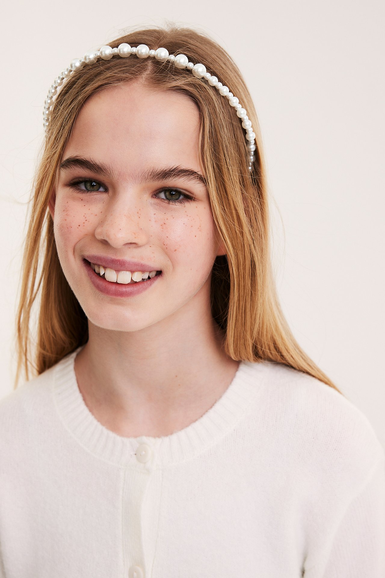 Forfra, et barn med et kremhvitt perlediadem og en kremfarget knappet cardigan.