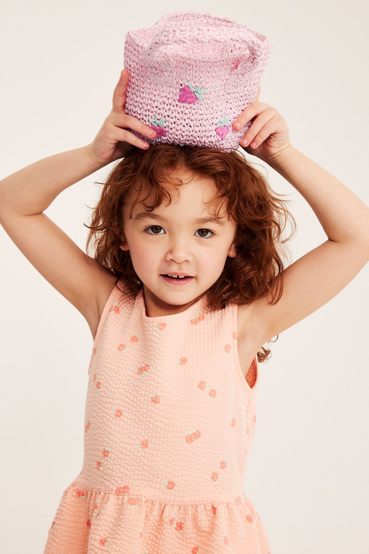 Edestä: Lapsi pitelee vaaleanpunaista olkilaukkua mansikkakirjailulla päällään, yllään persikkamekko.