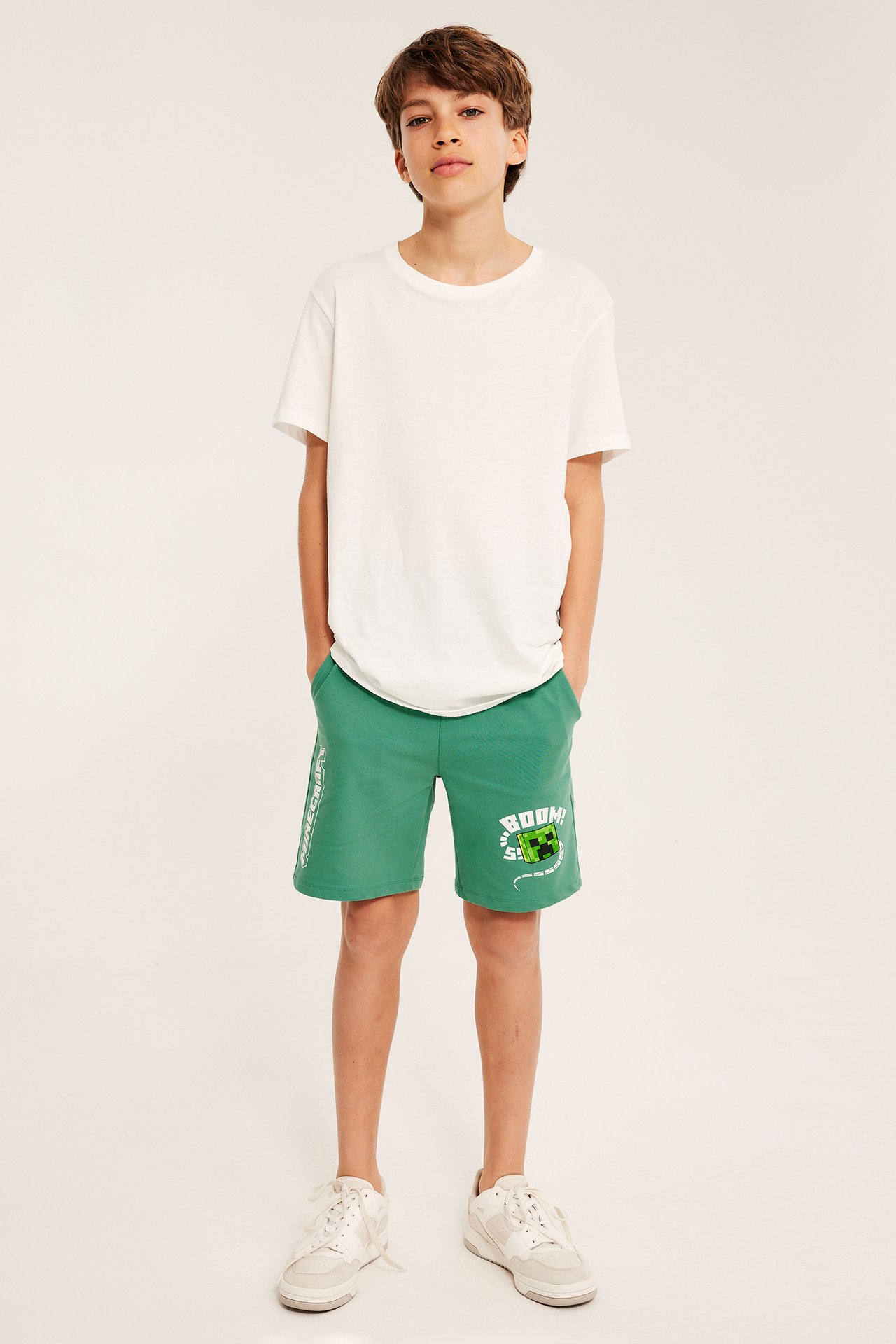 Z przodu: dziecko w zielonych szortach dresowych Minecraft, białej koszulce i białych sneakersach.