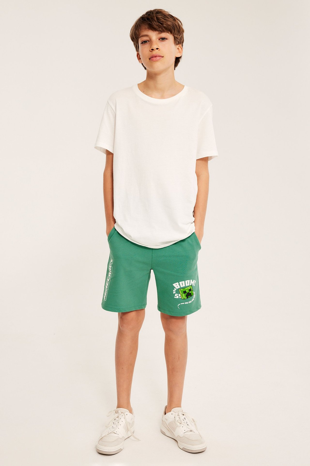 Z przodu: dziecko w zielonych szortach dresowych Minecraft, białej koszulce i białych sneakersach.