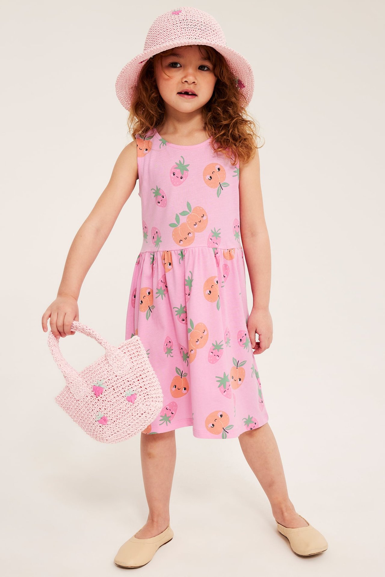 Edestä: Lapsi vaaleanpunaisessa hedelmäkuvioisessa mekossa, vaaleanpunainen olkihattu, pitäen vaaleanpunaista olkilaukkua, beiget ballerinat.