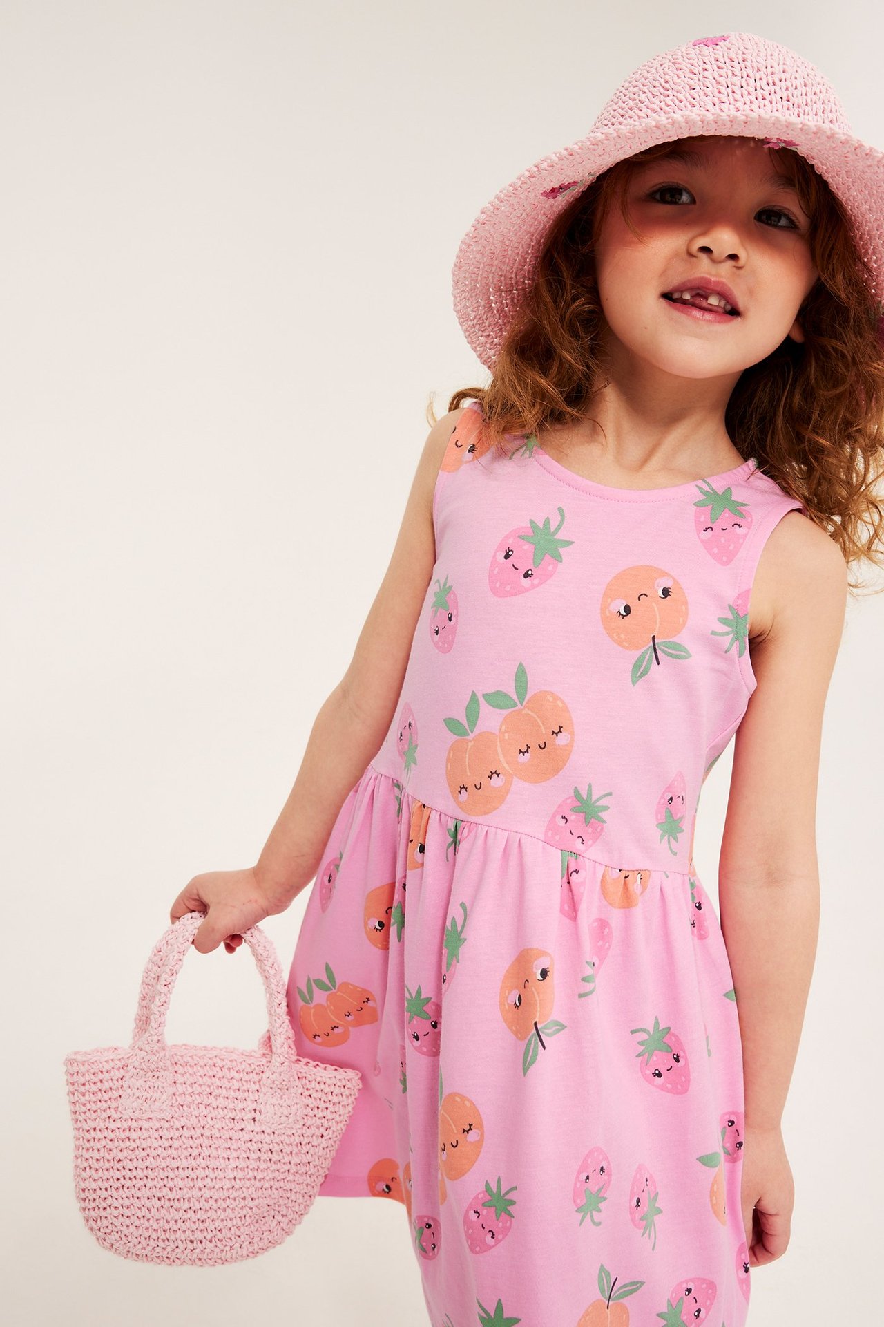 Edestä: Tyttö vaaleanpunaisessa hedelmäkuvioisessa puuvillamekossa, aurinkohatussa ja neulotussa laukussa.
