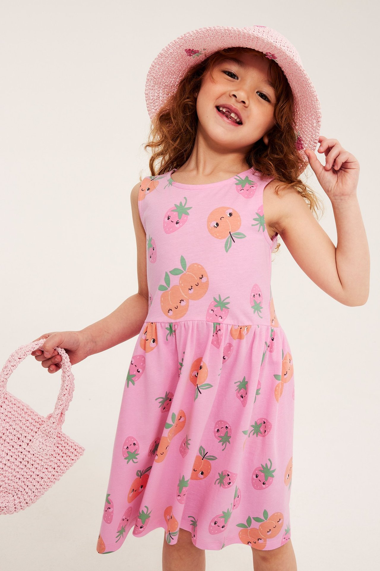 Widok z przodu: Dziecko w jasnoróżowej bawełnianej sukience z brzoskwiniowo-truskawkowym wzorem, różowy kapelusz, trzyma torebkę.
