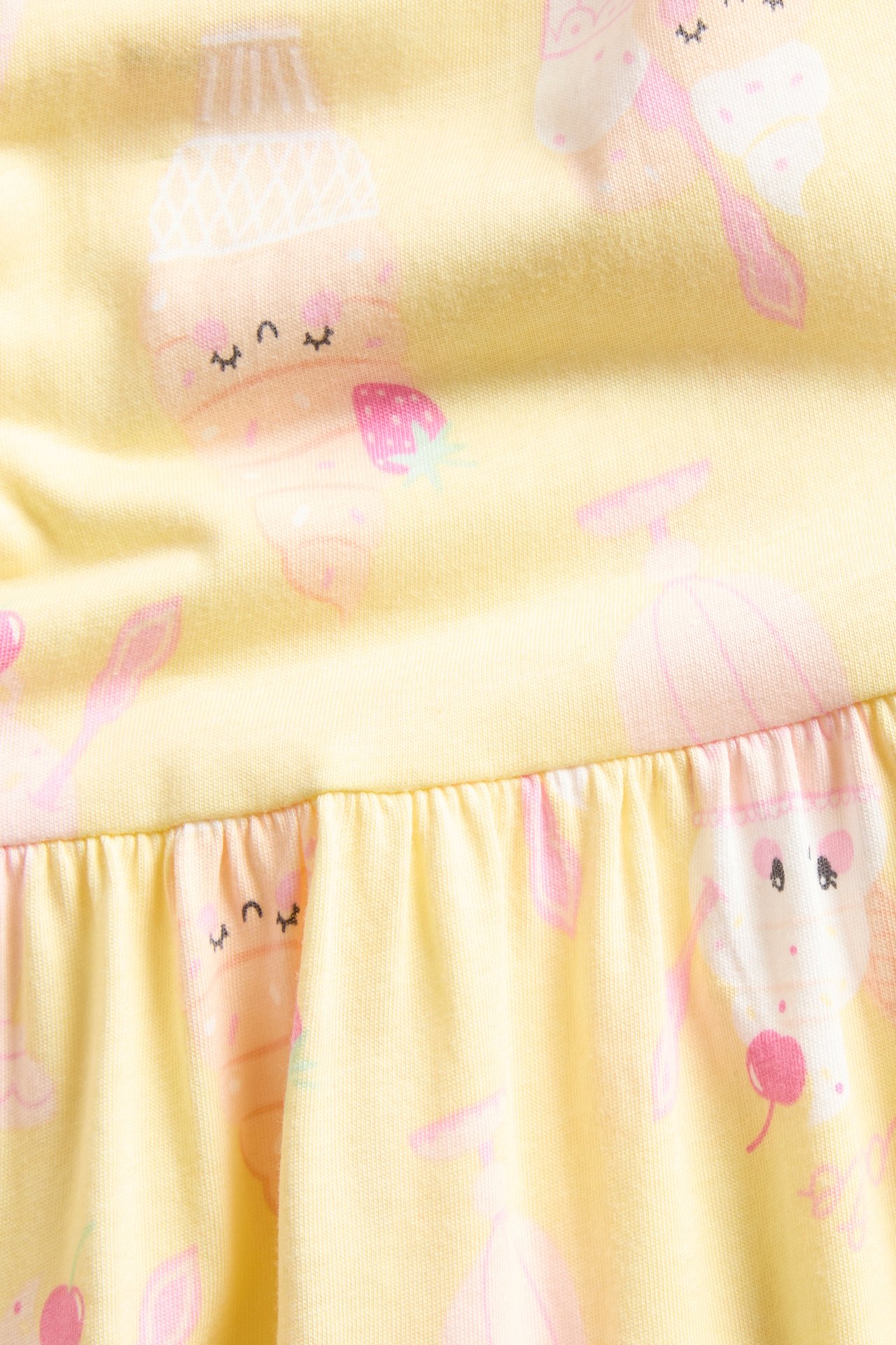 Zbliżenie na jasnożółtą bawełnianą sukienkę dziecięcą z różowymi wzorami lodów i truskawek oraz marszczonym dołem.