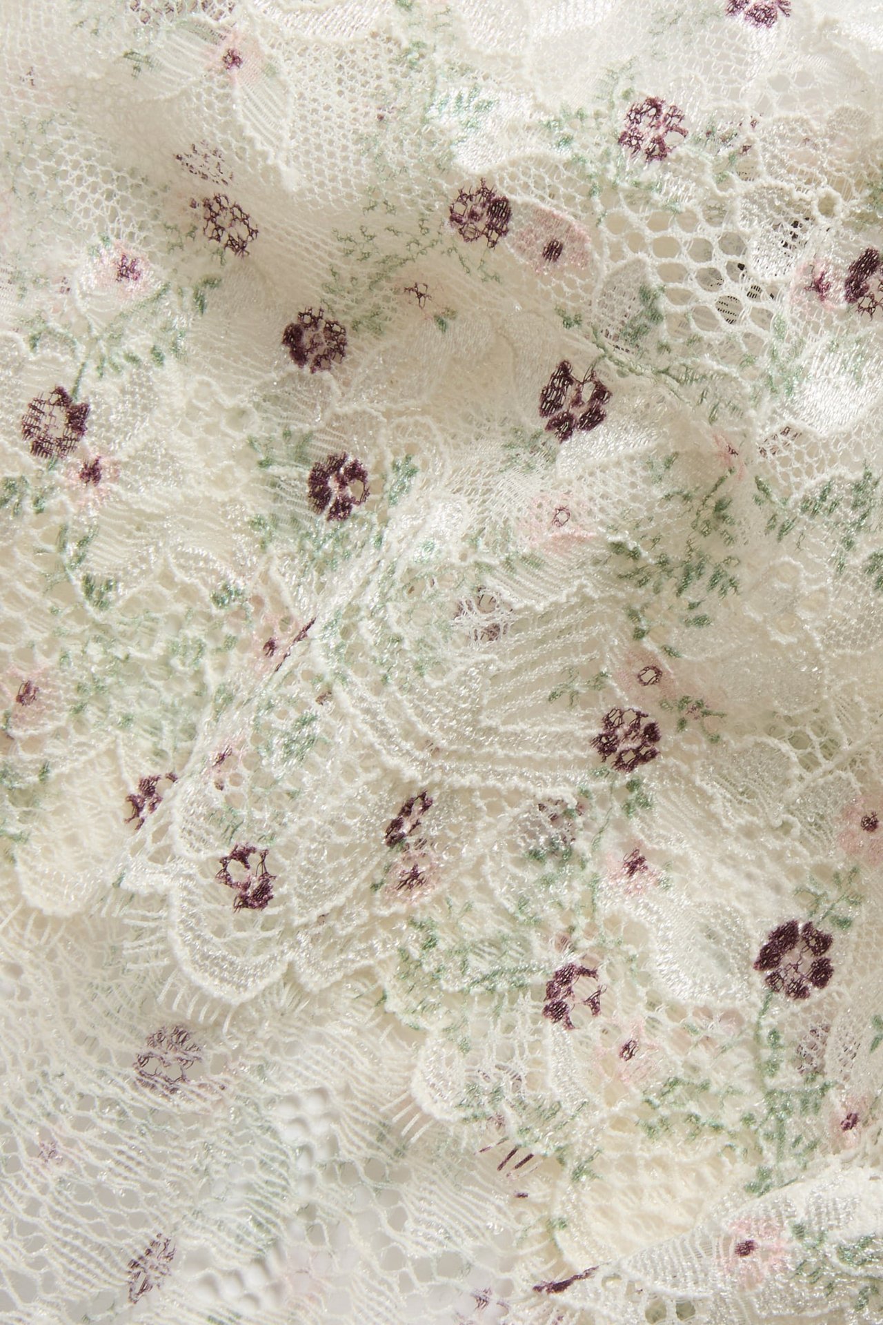 Närbild av vit spetstyg med broderade lila och rosa blommor samt gröna blad.