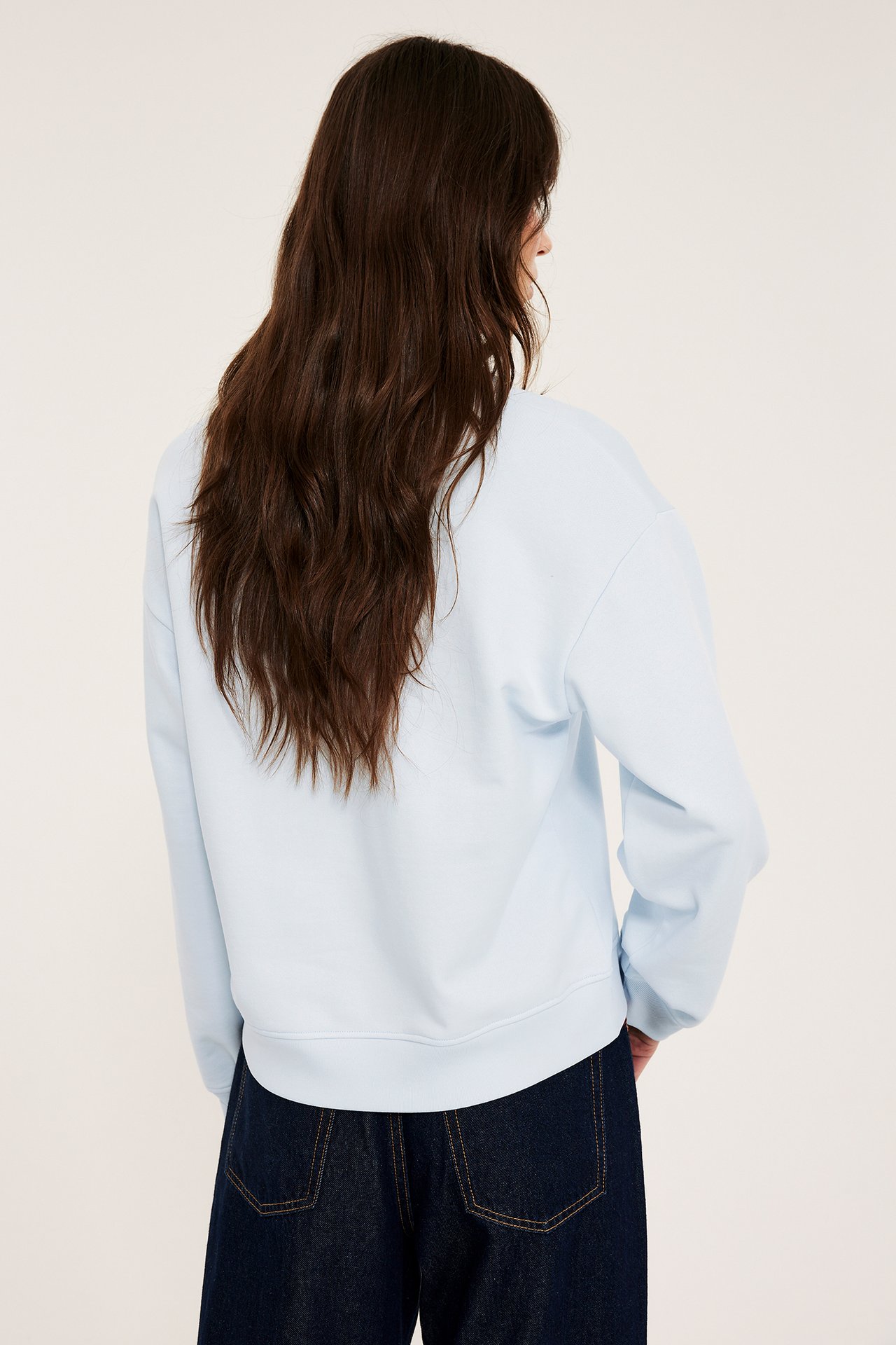 Nainen takaa, yllään vaaleansininen puuvillacollegepaita ja tummansiniset farkut.