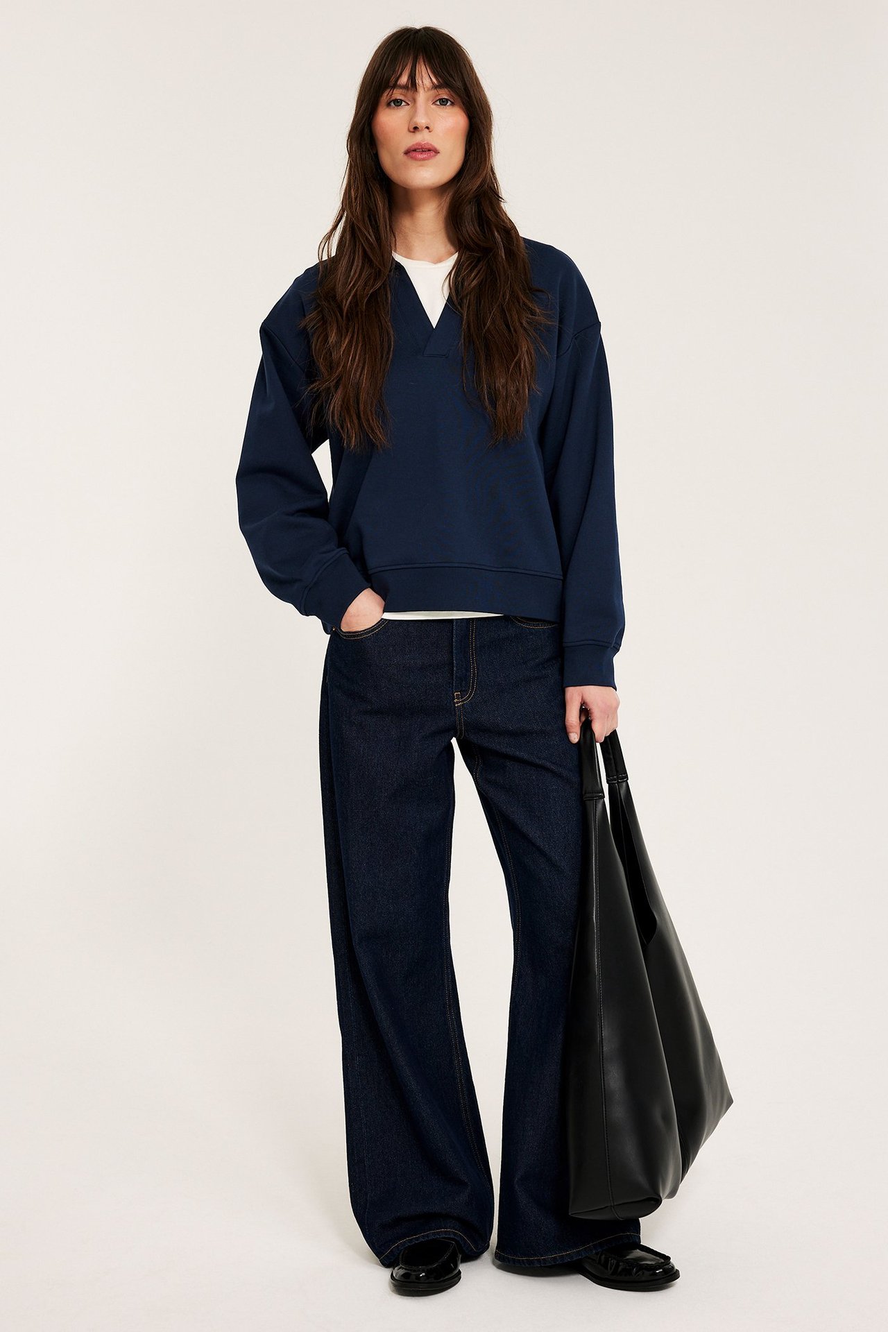 Edestä: Nainen tummansinisessä collegepaidassa, valkoisen t-paidan päällä, tummissa farkuissa, musta laukku.